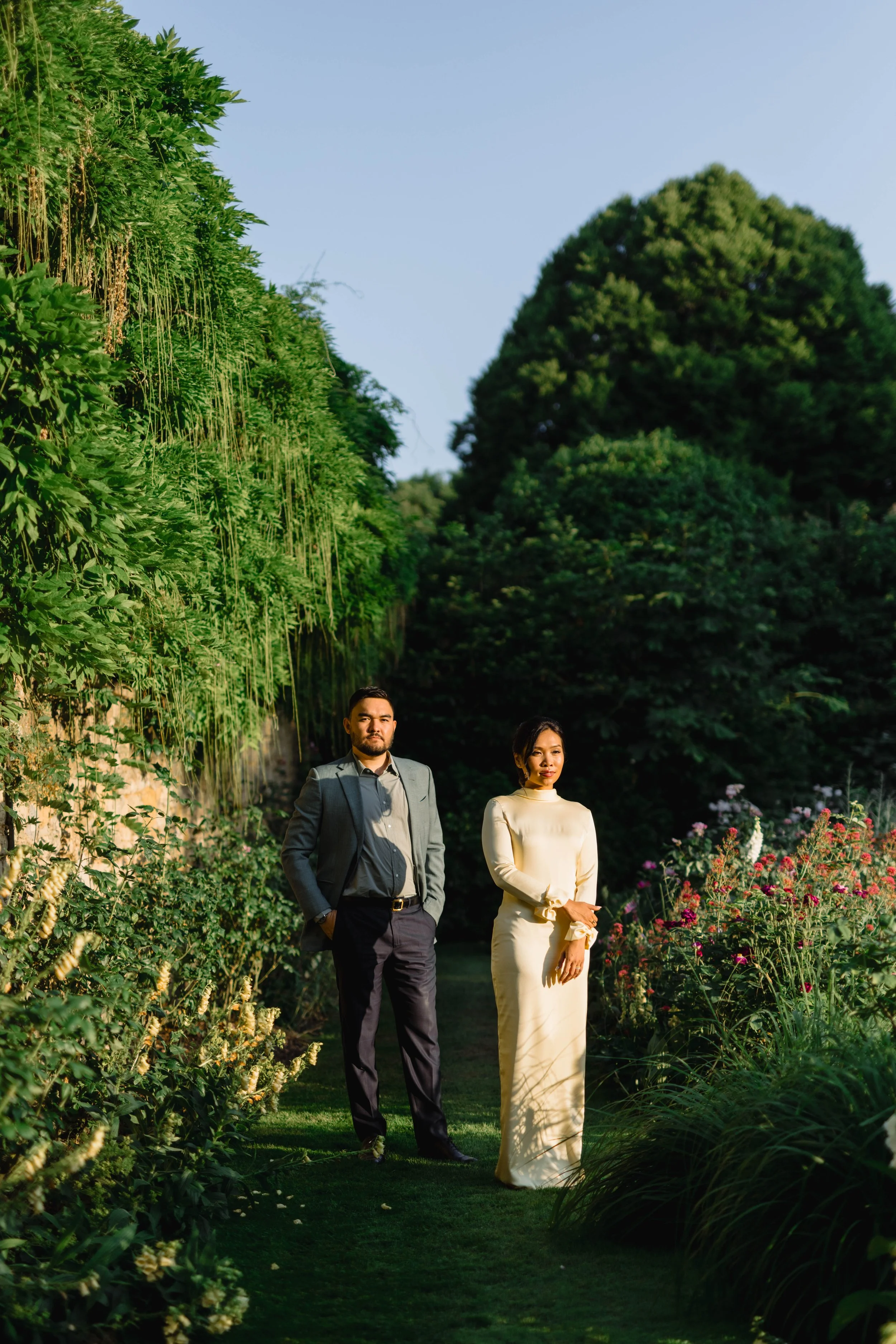 A man in a suit and a woman in a cream-colored dress standing on a garden path surrounded by lush greenery and flowers.
