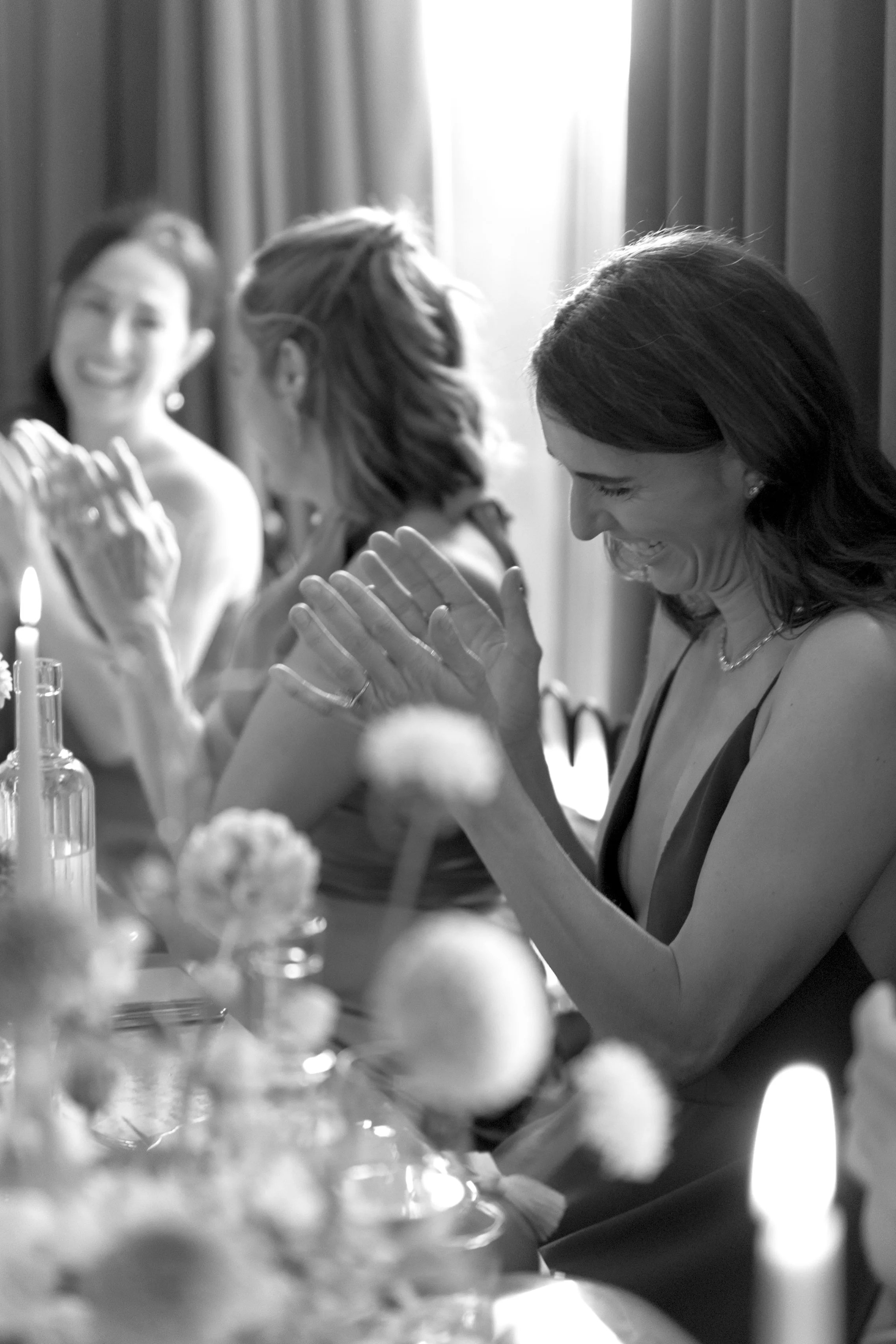 Women sitting together at a table, clapping and smiling, celebrating a special occasion in a well-lit room with flowers and candles.