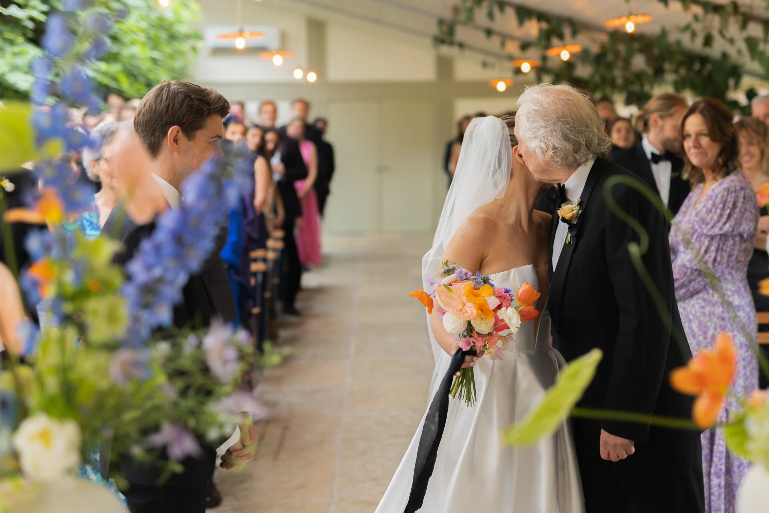 A bride and an older man, likely her father, sharing a kiss at her wedding, surrounded by wedding guests in an indoor venue decorated with flowers.