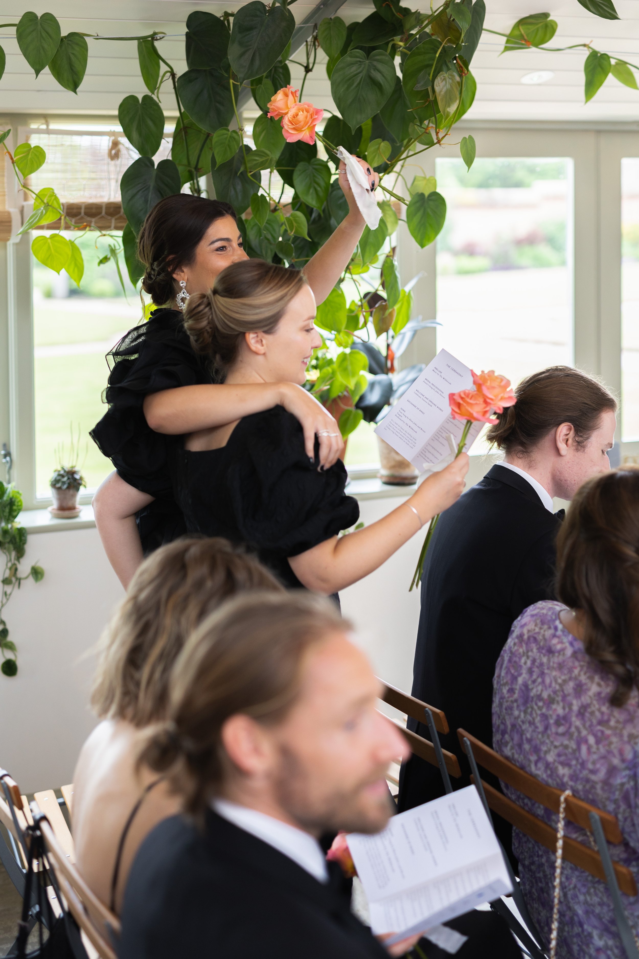A woman with dark hair and earrings is standing, holding a bouquet of peach roses and a program, while a young girl with blonde hair and a black dress sits on her shoulders, also holding a peach rose, during a wedding ceremony. Others seated are read