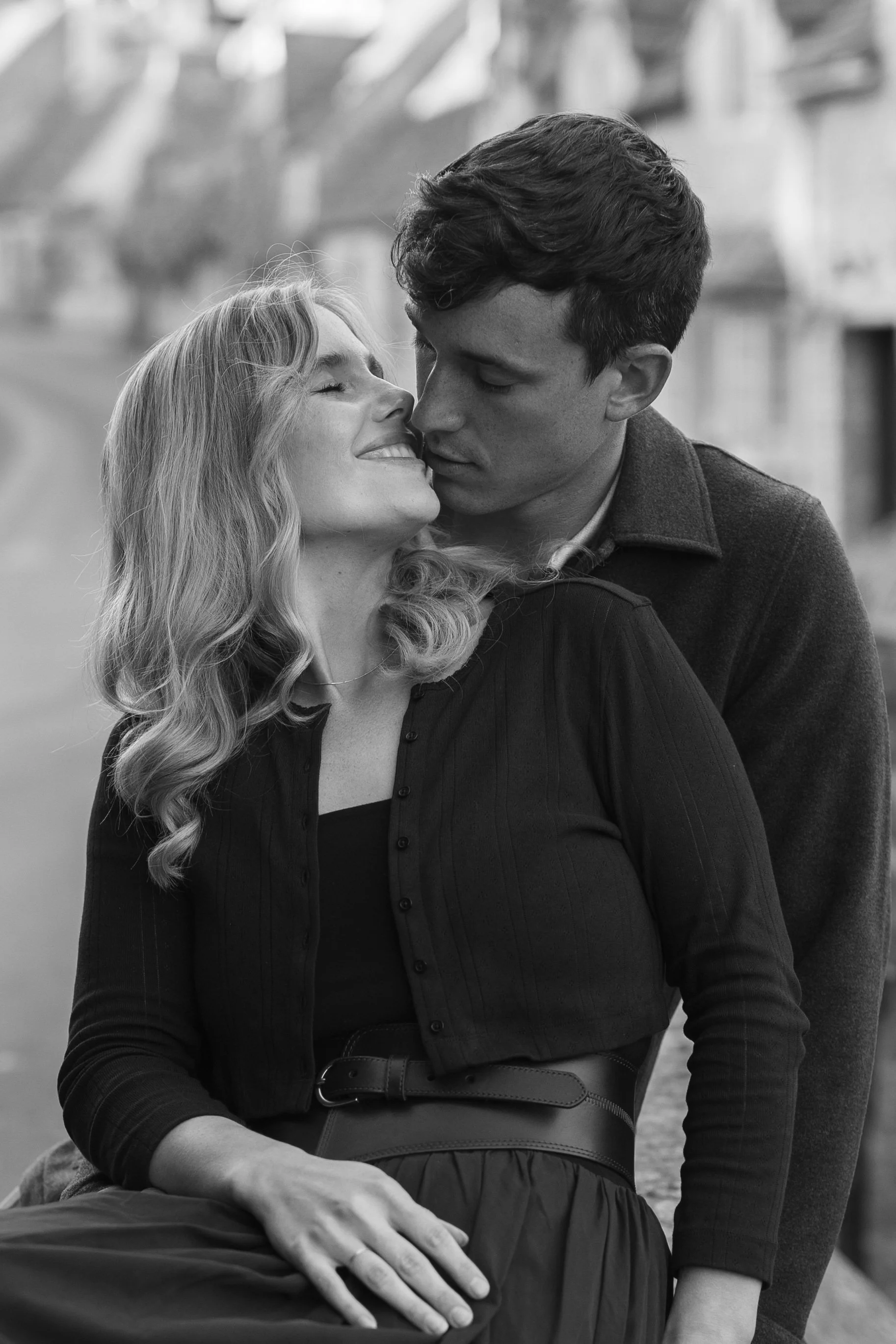 A black and white photo of a smiling woman with wavy hair and a man with short hair close together, appearing intimate, outdoors with blurred background.
