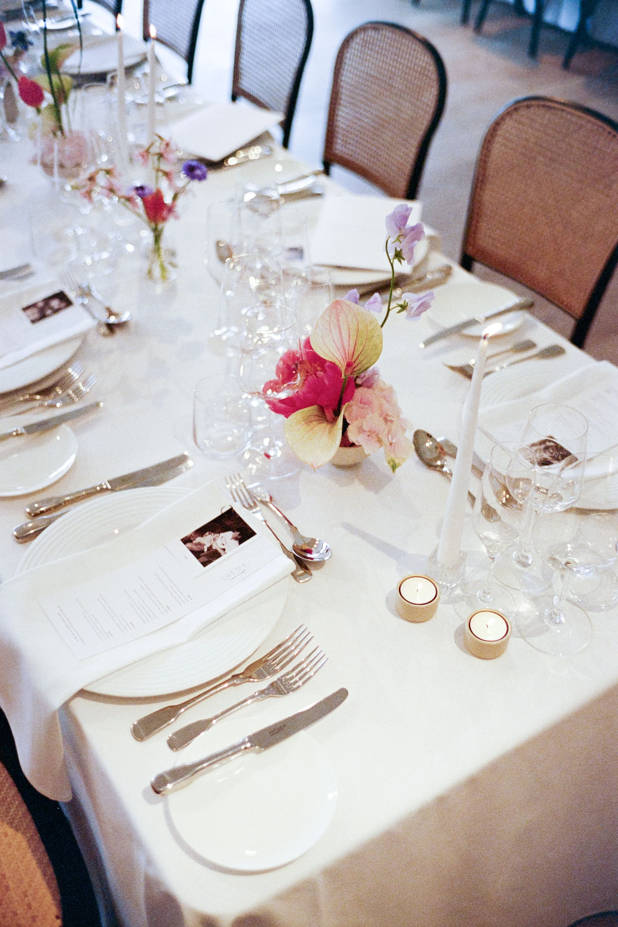 A formal dining table decorated with pink and purple flowers, wine glasses, candles, and place settings, including white plates, silver cutlery, and printed menus.