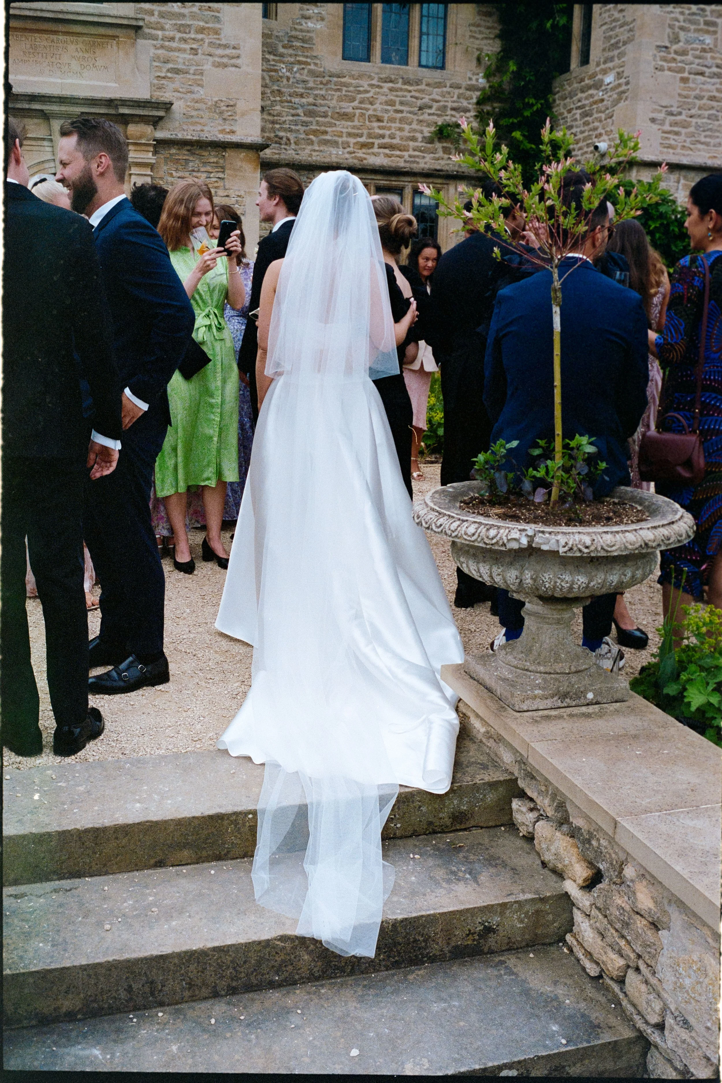 A bride in a white wedding dress and veil standing on stone steps outdoors, surrounded by wedding guests in formal attire, near a historic stone building with ivy growing on its walls.