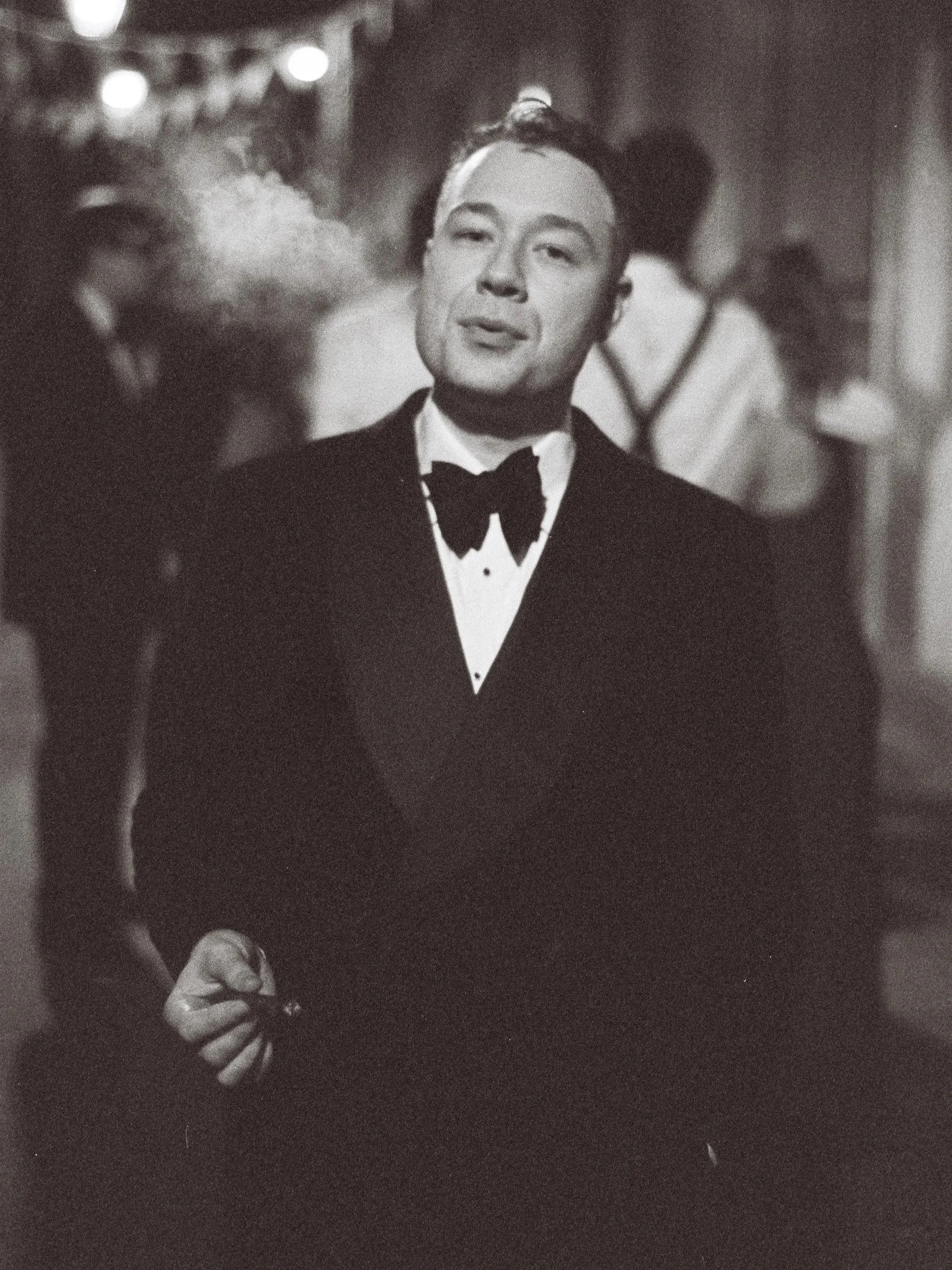 A man in a tuxedo holding a cigarette in his hand at a wedding in a black and white 35mm photograph.