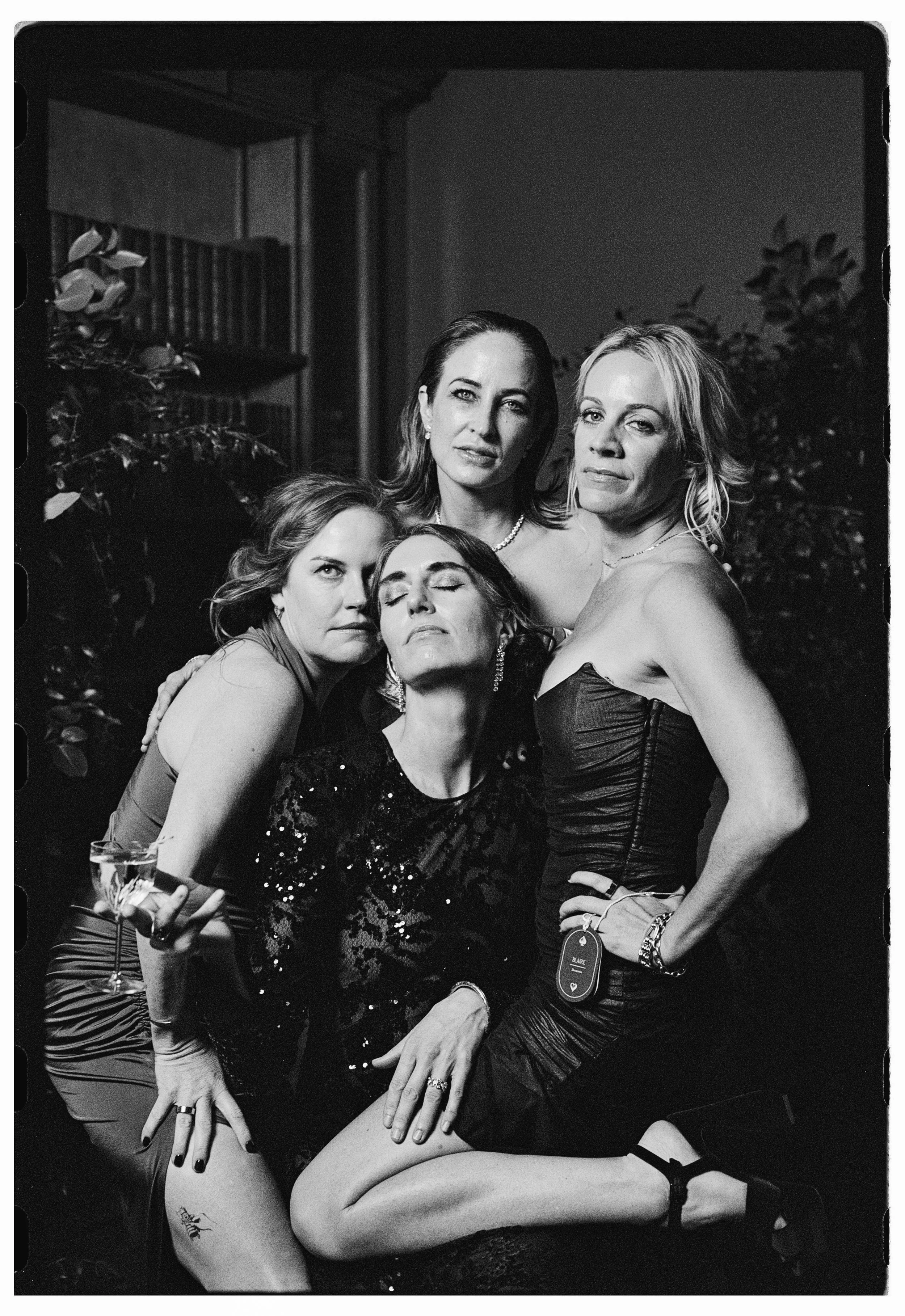 Black and white photograph of four women dressed in elegant evening wear posing together at a wedding. One woman is holding a glass of wine, and they appear to be enjoying themselves.