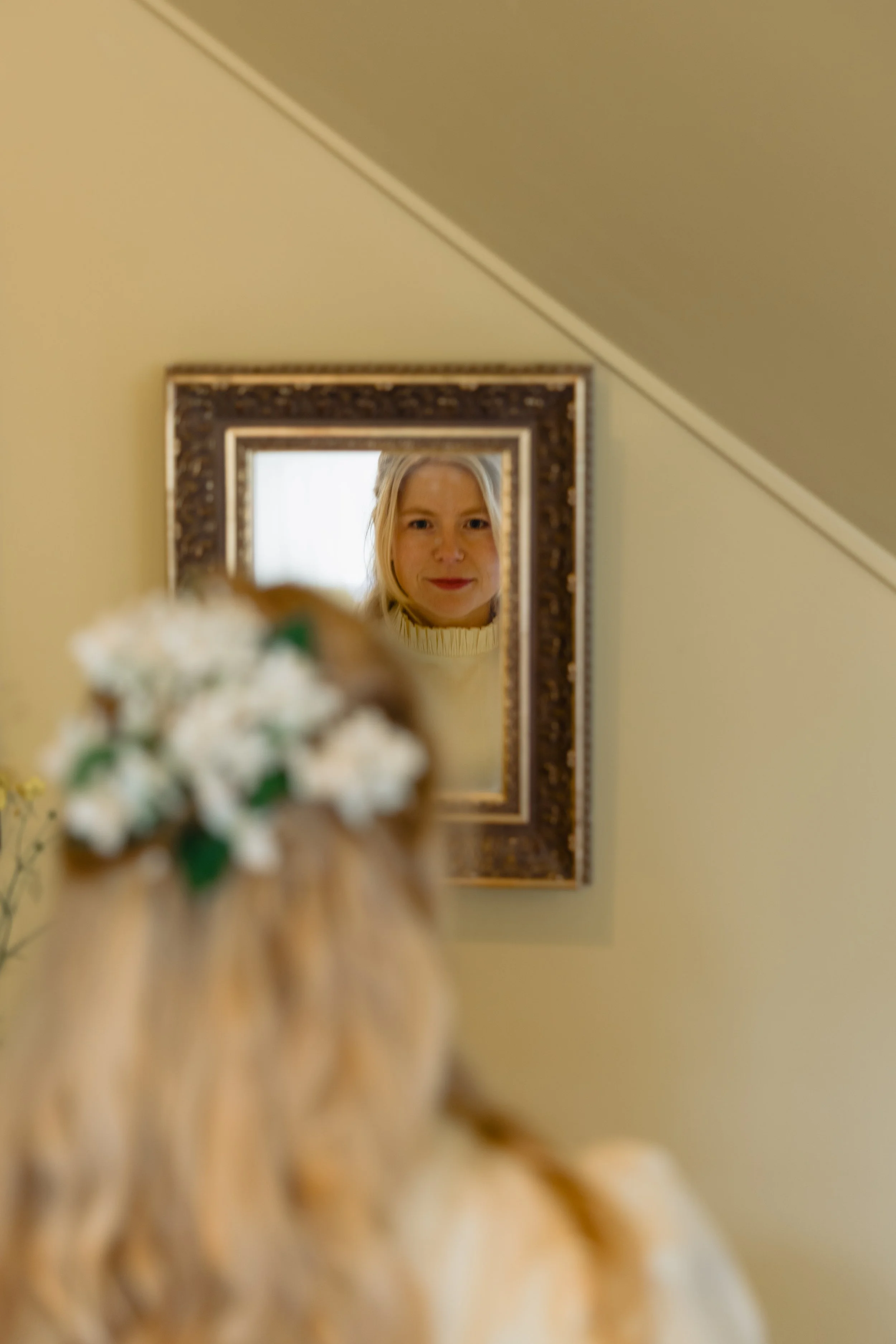 A woman with blonde hair looking into a mirror, with her face reflected, on a wall behind a blurred bouquet of white flowers.