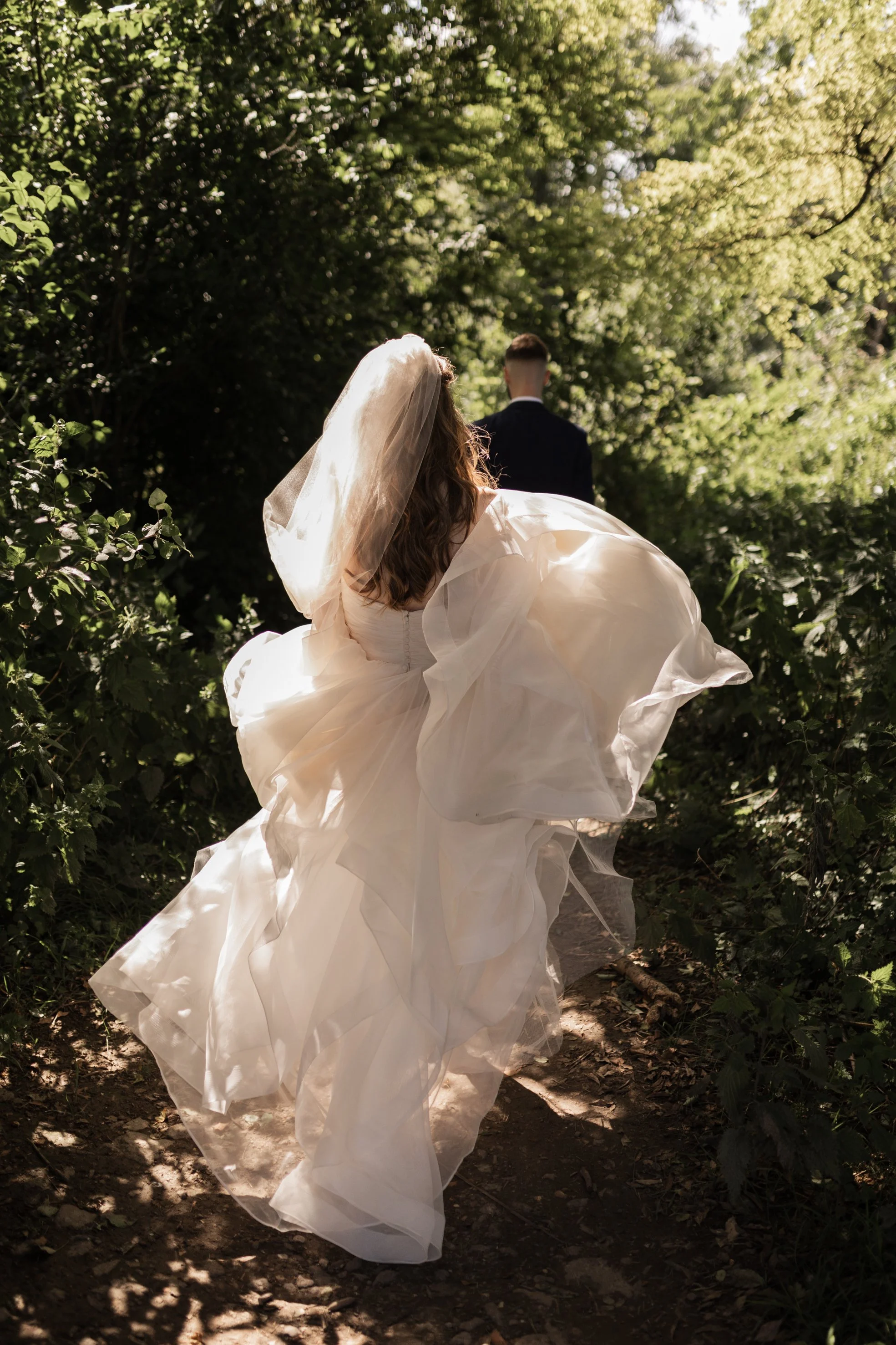 Bride in a white wedding dress and veil walking along a wooded path with groom in the background.