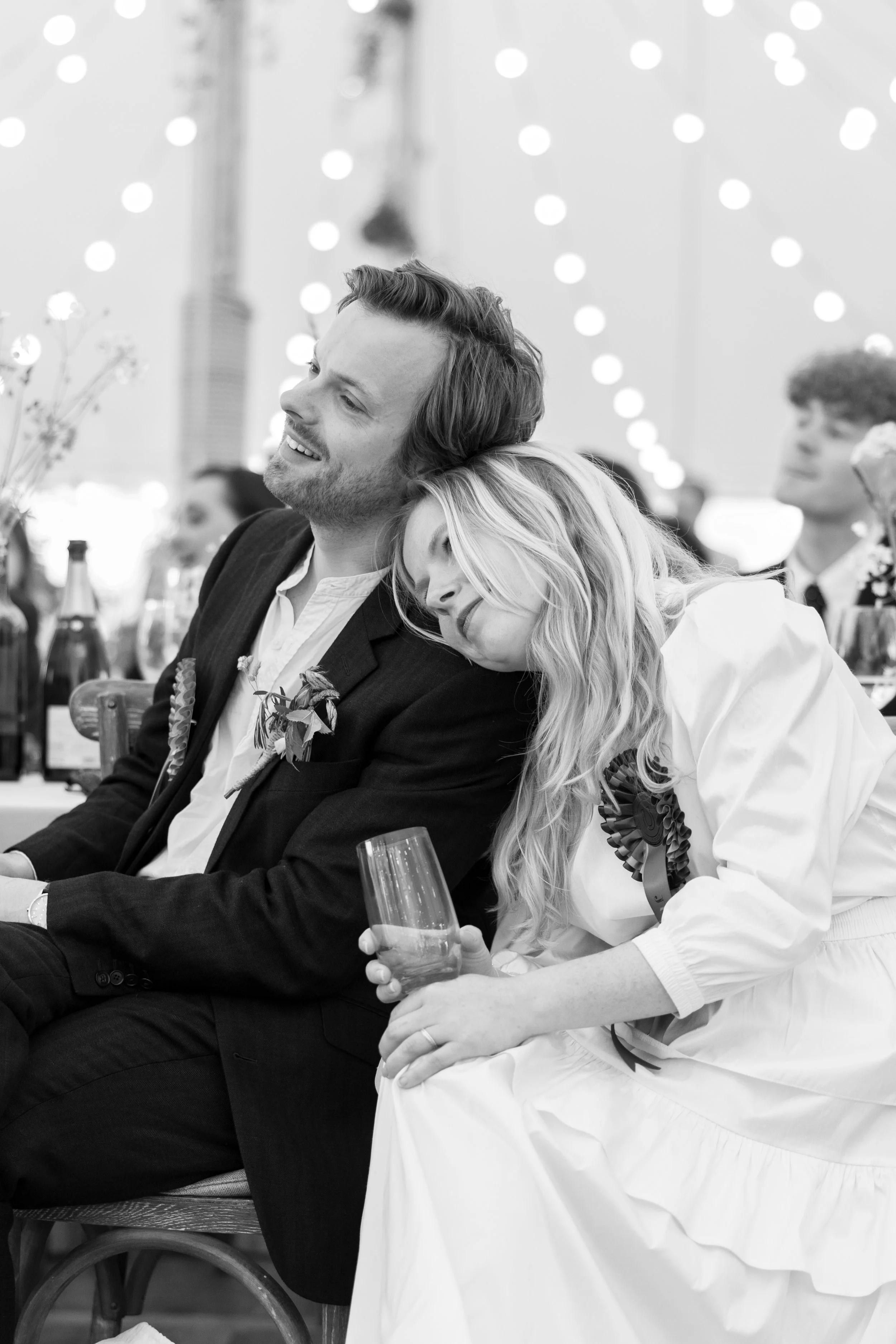 A black and white photo of a couple sitting close together at a celebration, with the woman resting her head on the man's shoulder. They are dressed formally, with the woman holding a wine glass and both smiling softly. In the background, there are o