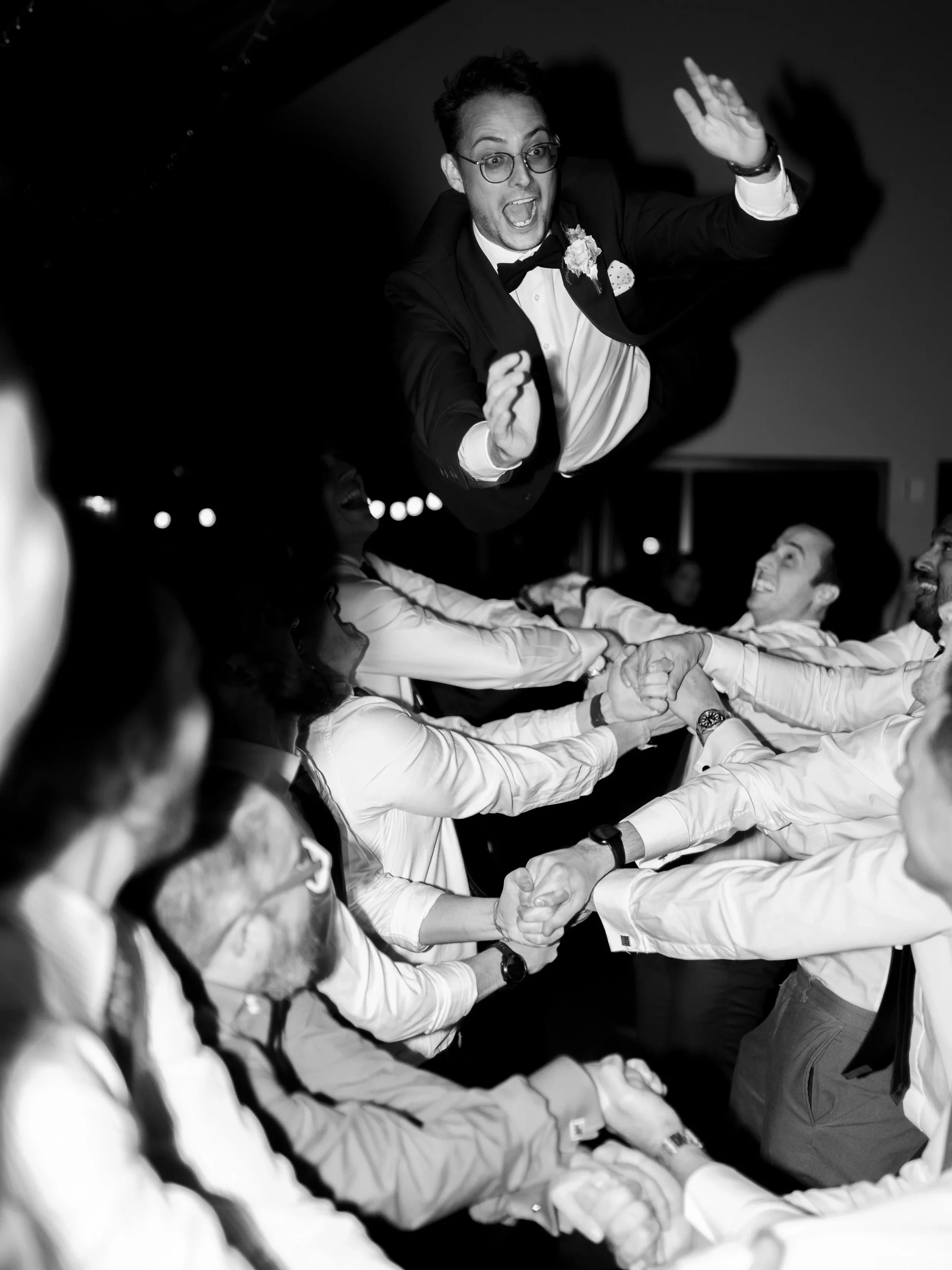 Man in tuxedo and glasses being thrown into the air at a wedding celebration with friends.
