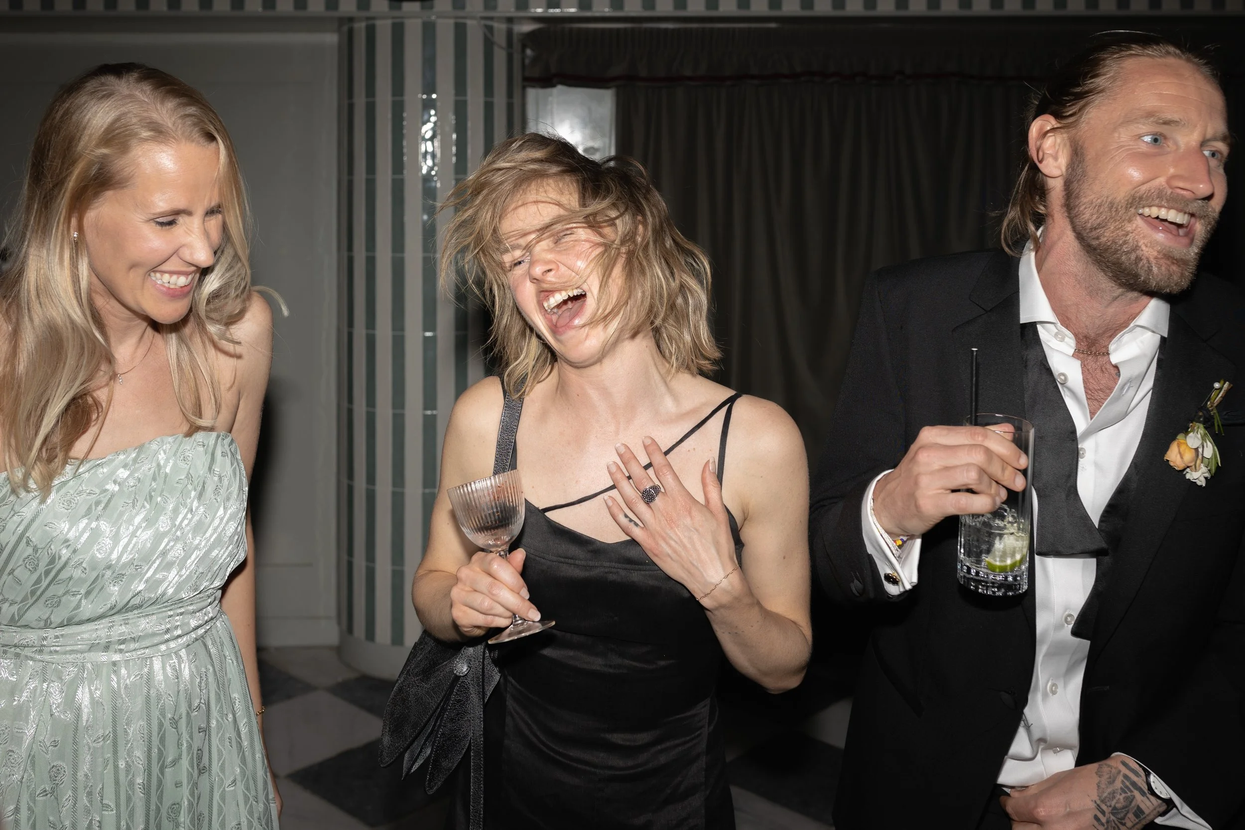 Three people dressed in formal attire celebrating and laughing, holding drinks at a social event.