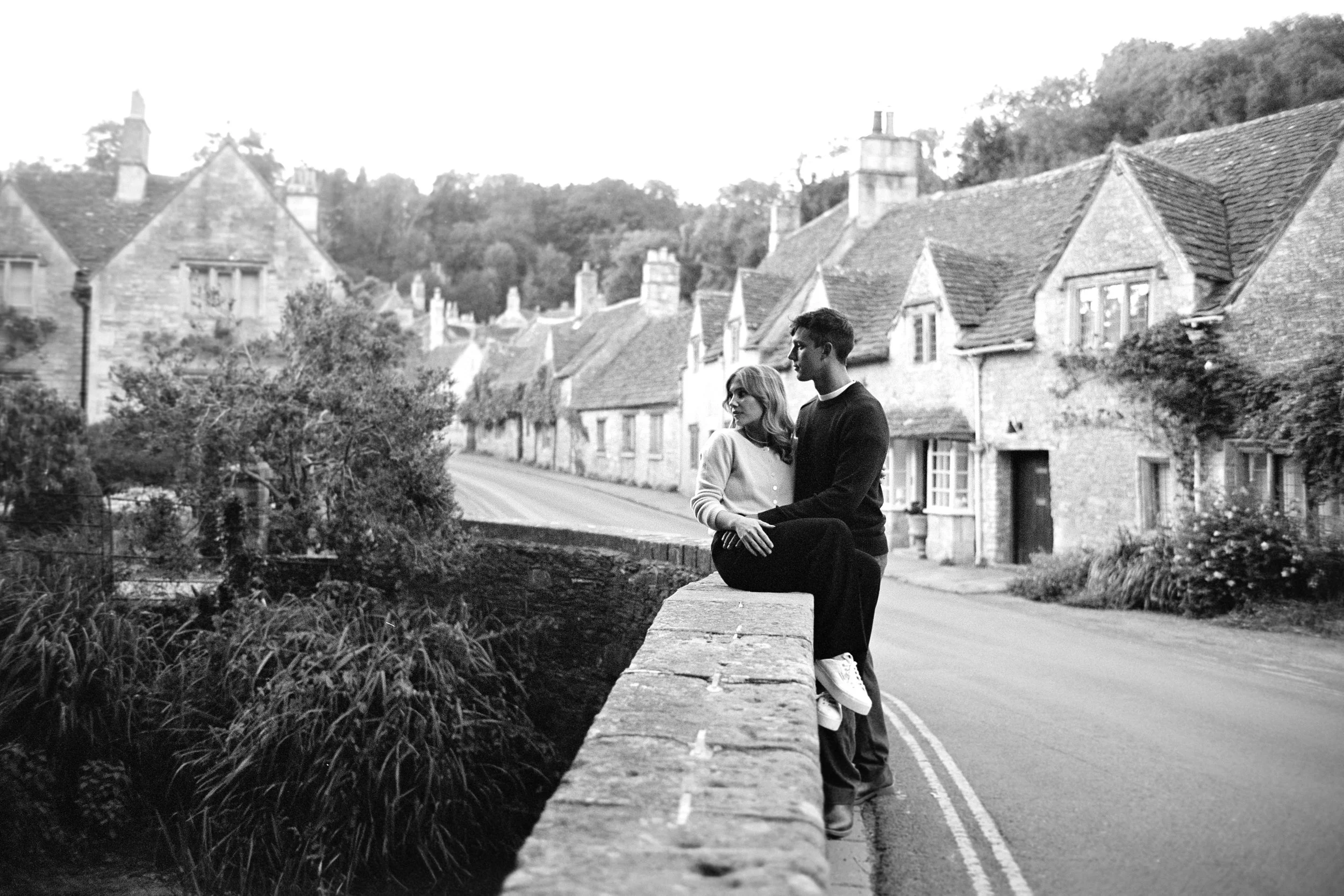 A black-and-white photo of a young couple sitting closely on a stone wall by a street in a quaint village, with houses and trees in the background.