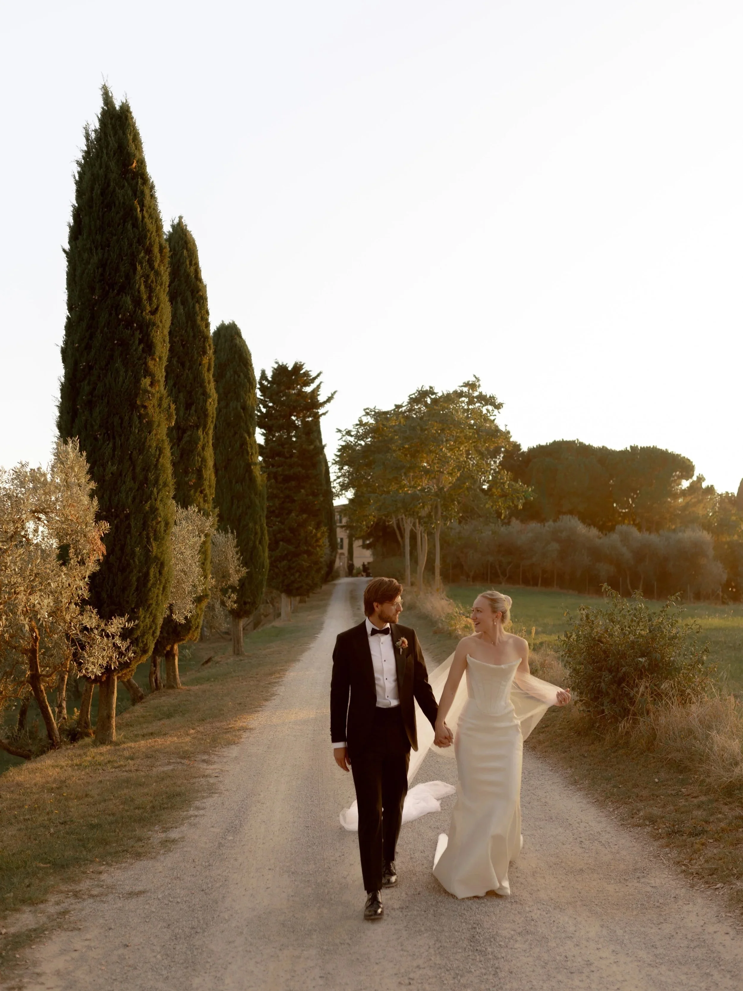 A bride and groom walking hand in hand on a dirt road surrounded by tall trees during sunset at their Tuscan wedding.