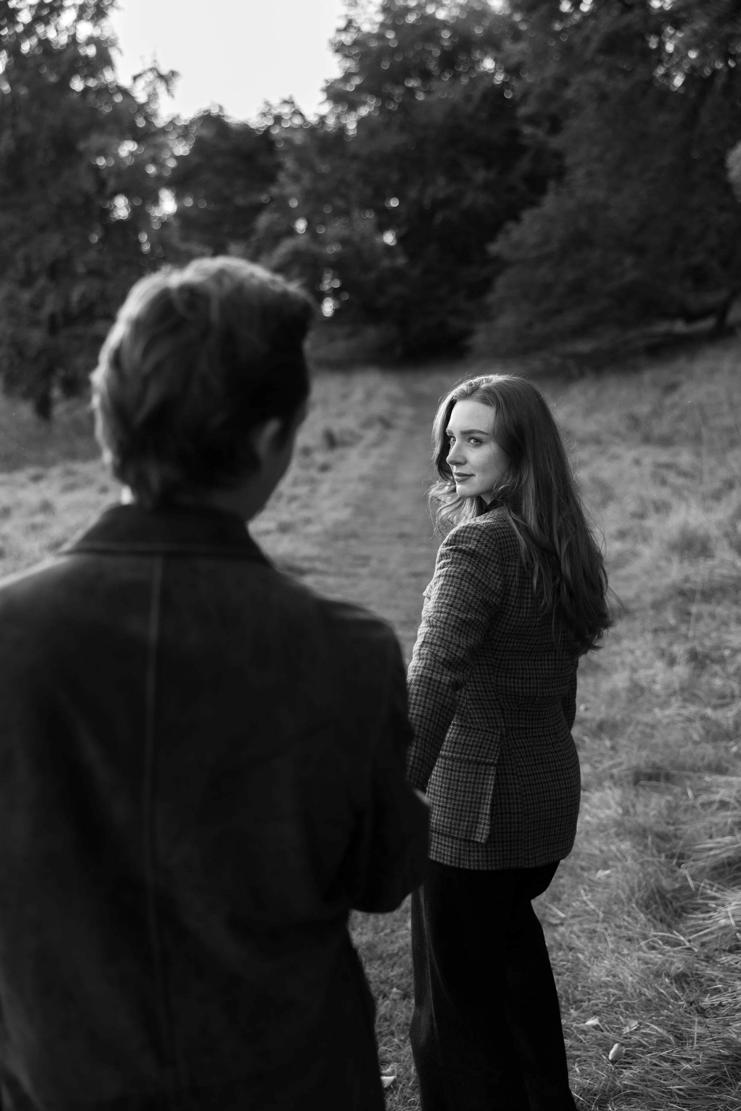 A woman with long hair and a checkered blazer looking over her shoulder outdoors, with a man in the foreground, blurred, in a natural setting with trees and grass.