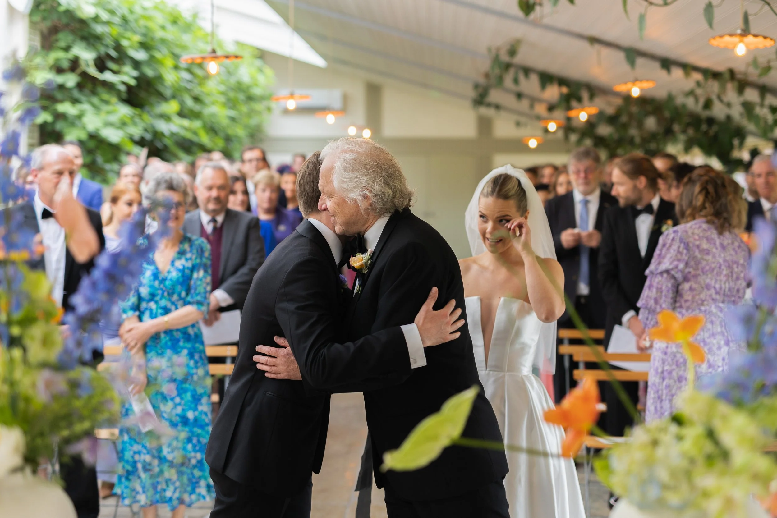 At a wedding reception, a groom and an older man are sharing a hug. The bride, wiping away tears, stands nearby with a smile. The scene is filled with guests in formal attire, in a decorated indoor venue surrounded by flowers and greenery.