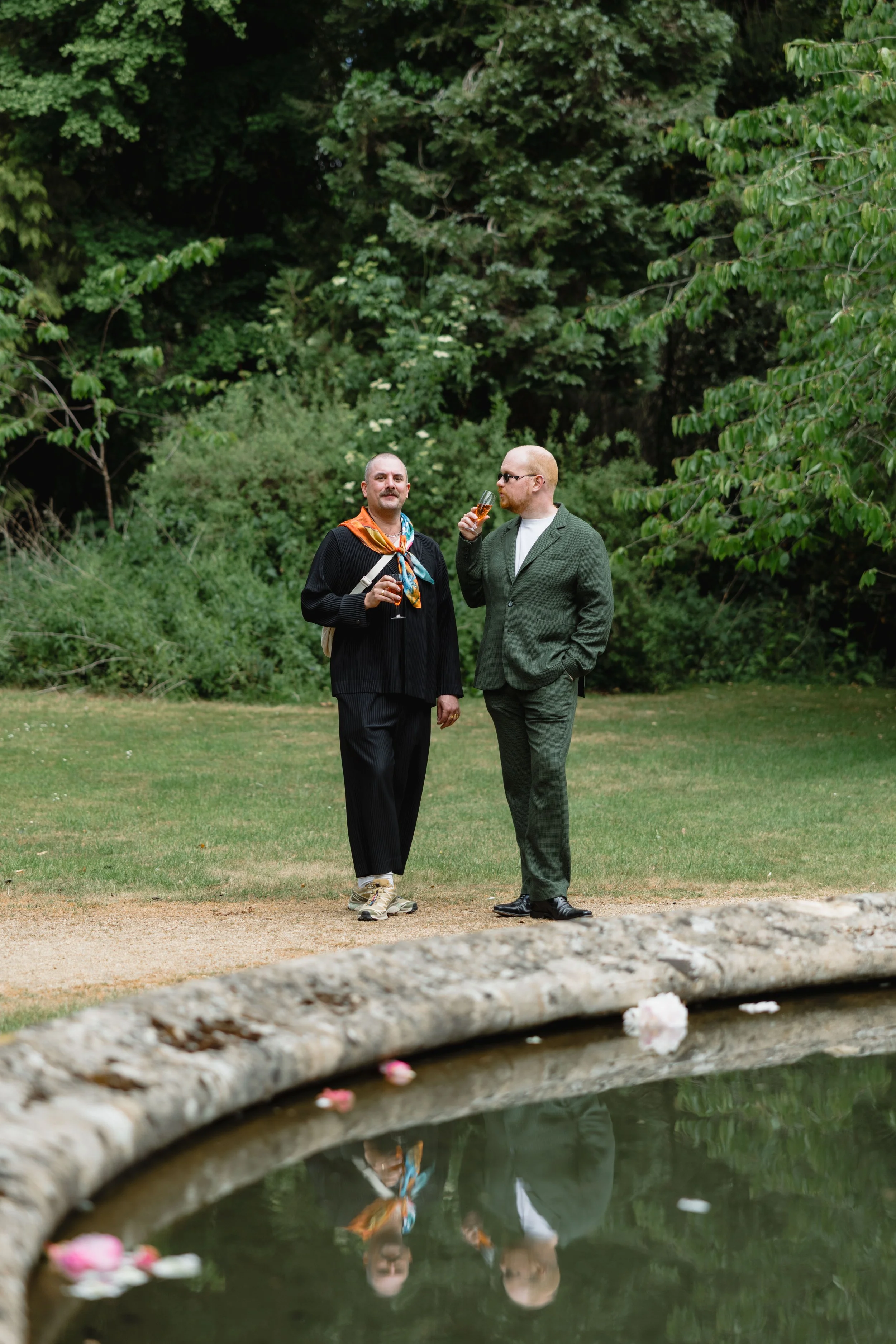 Two men in suits and casual shoes standing outdoors near a pond with floating flowers, holding glasses of drinks, talking amidst lush green trees and bushes.
