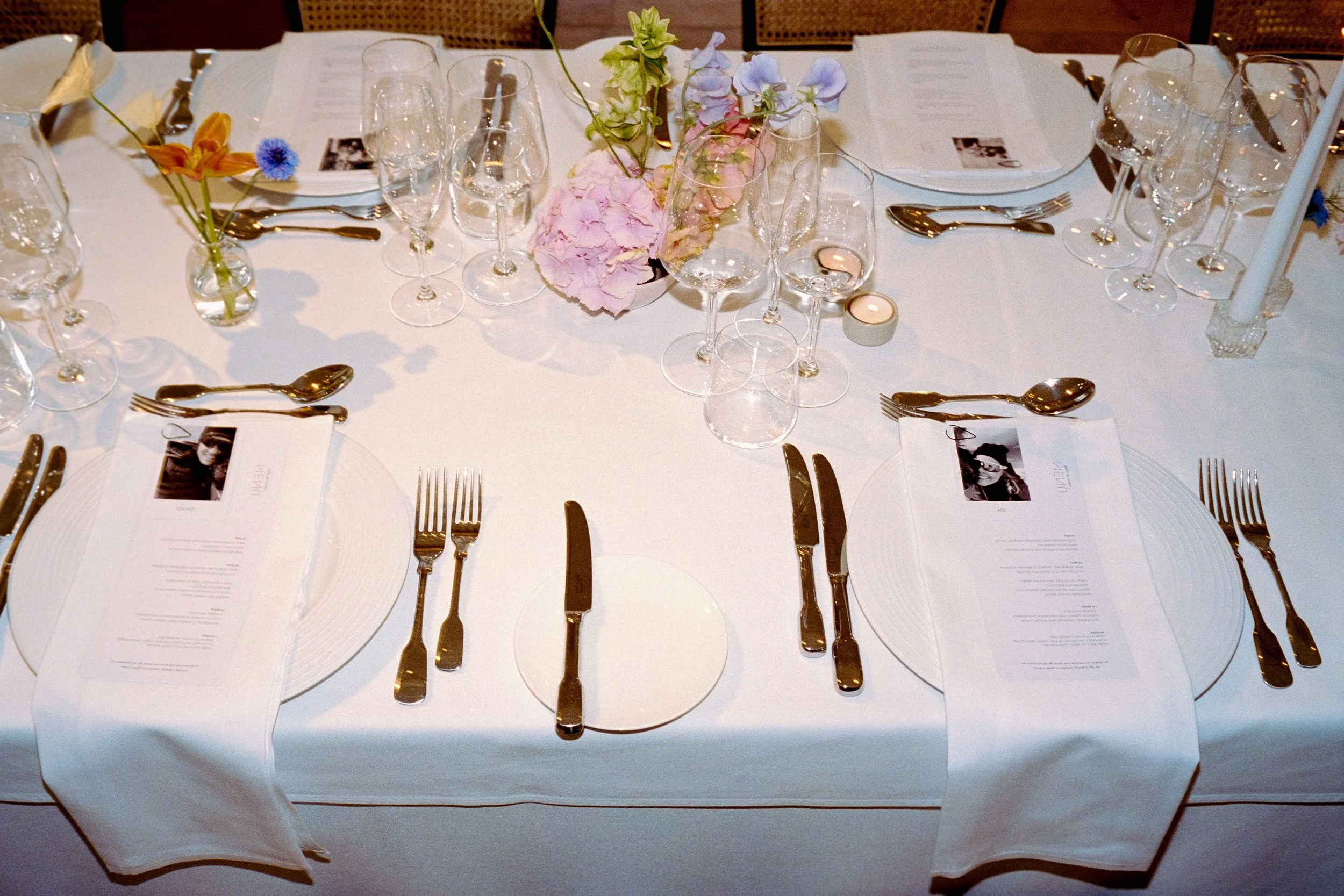 A formal dining table set with white tablecloth, multiple glasses, silverware, white napkins, pink and purple flowers in vases, and printed menus.