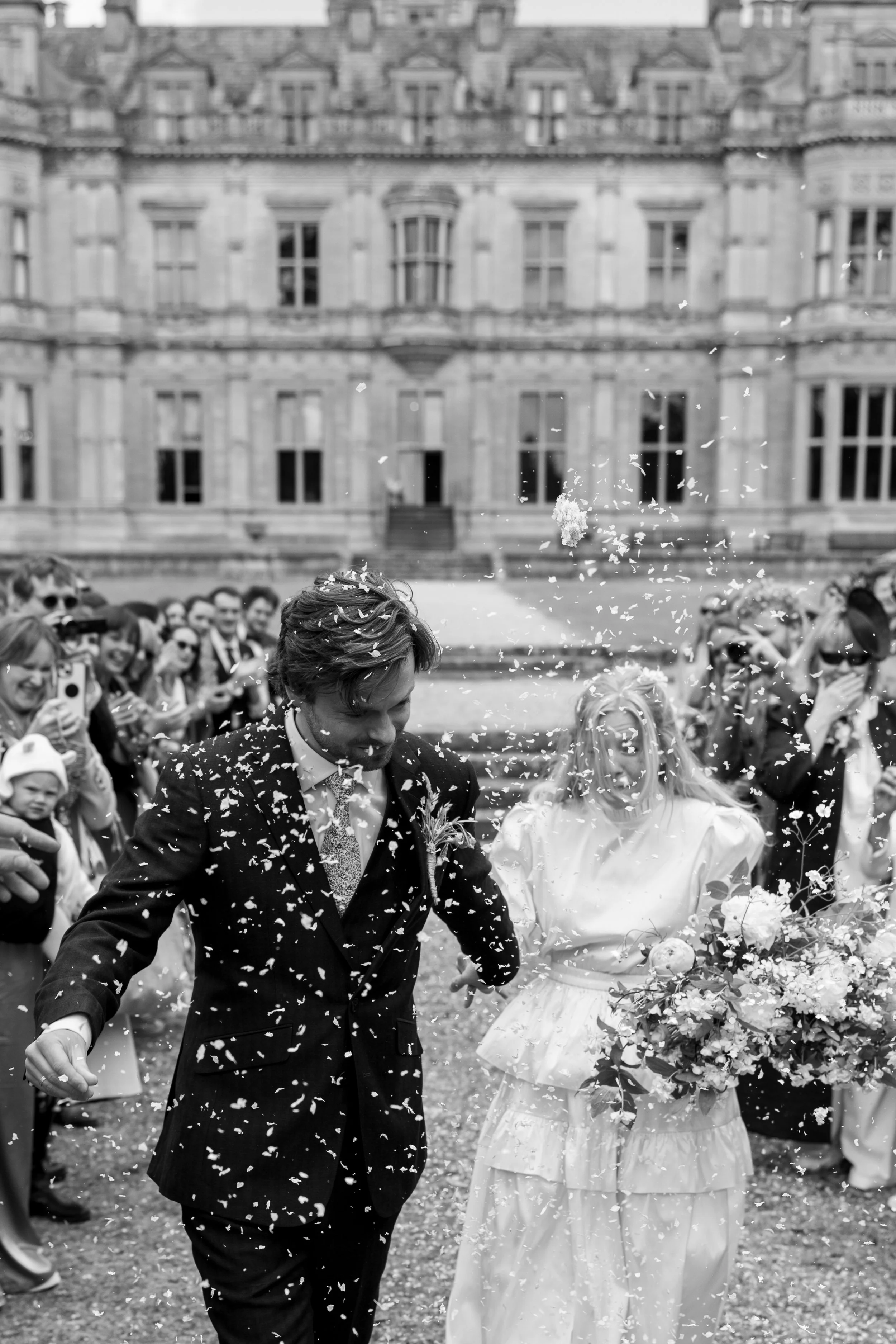 A black and white photograph of a newlywed couple walking outside a large, ornate building surrounded by friends and family, with confetti being thrown in the air.