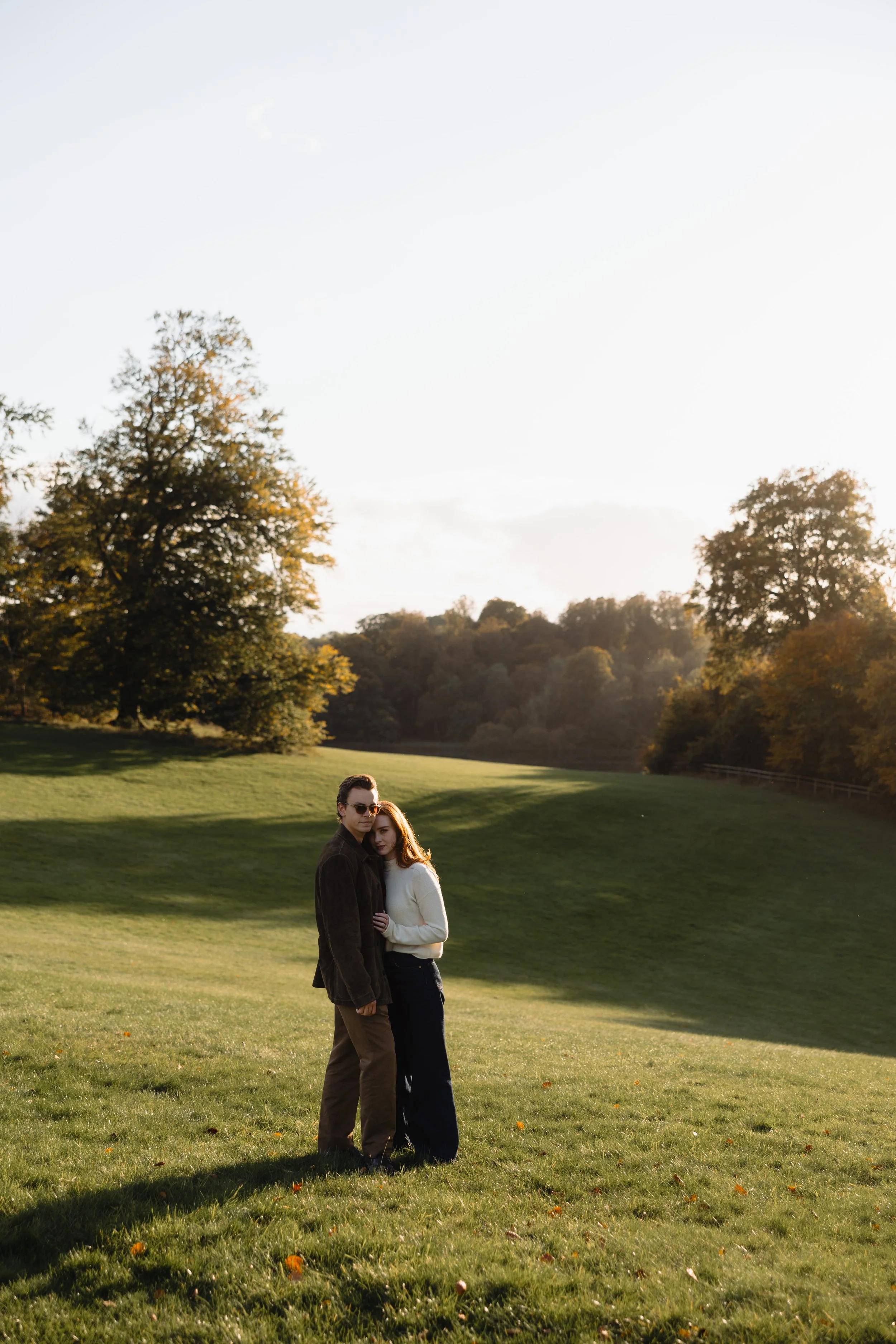 A couple stands close together in a grassy field during sunset with trees in the background.