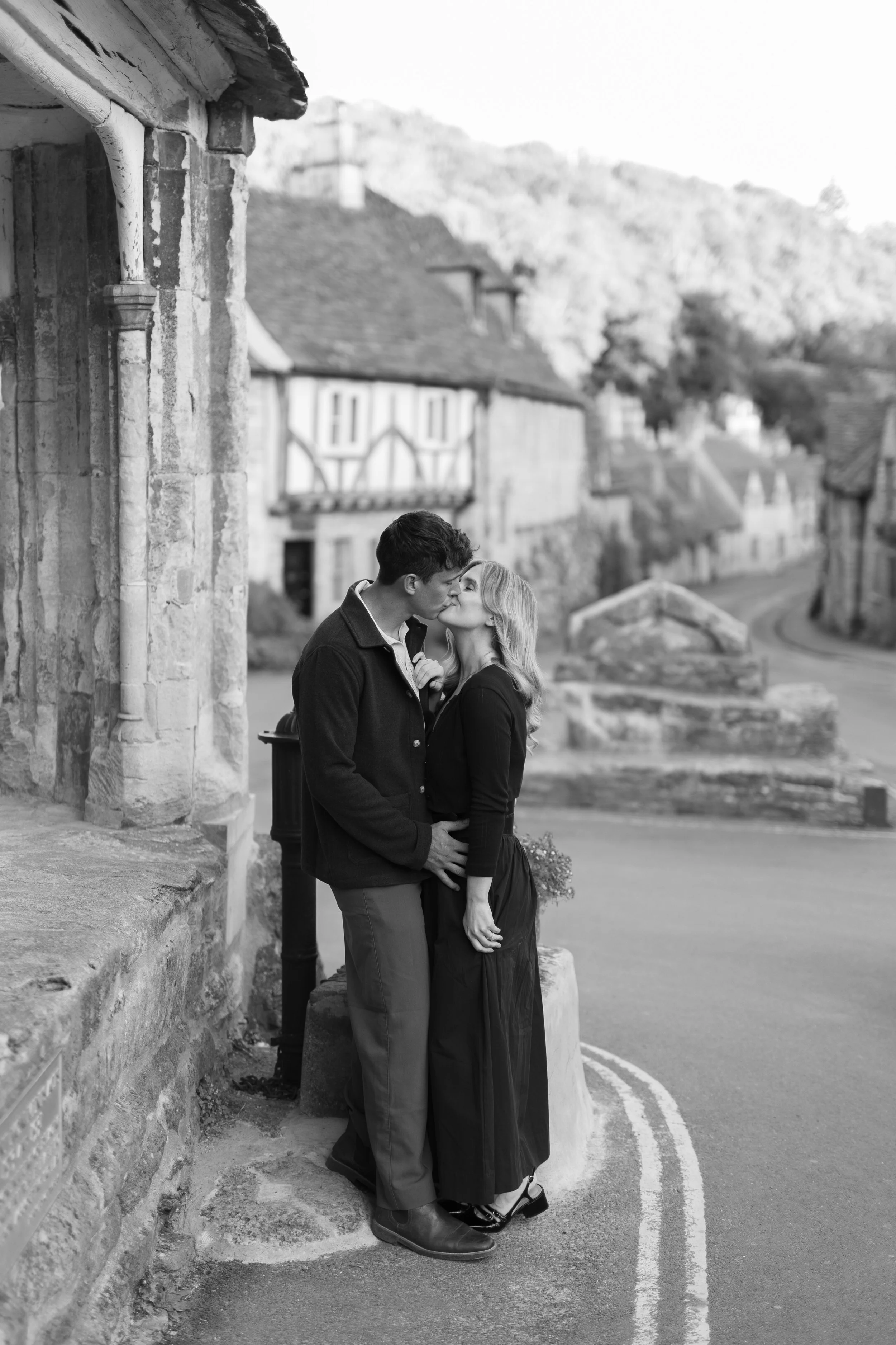 A black and white photo of a couple sharing a kiss on a quiet, historic street in a small town with old buildings and cobblestone, romantic atmosphere.