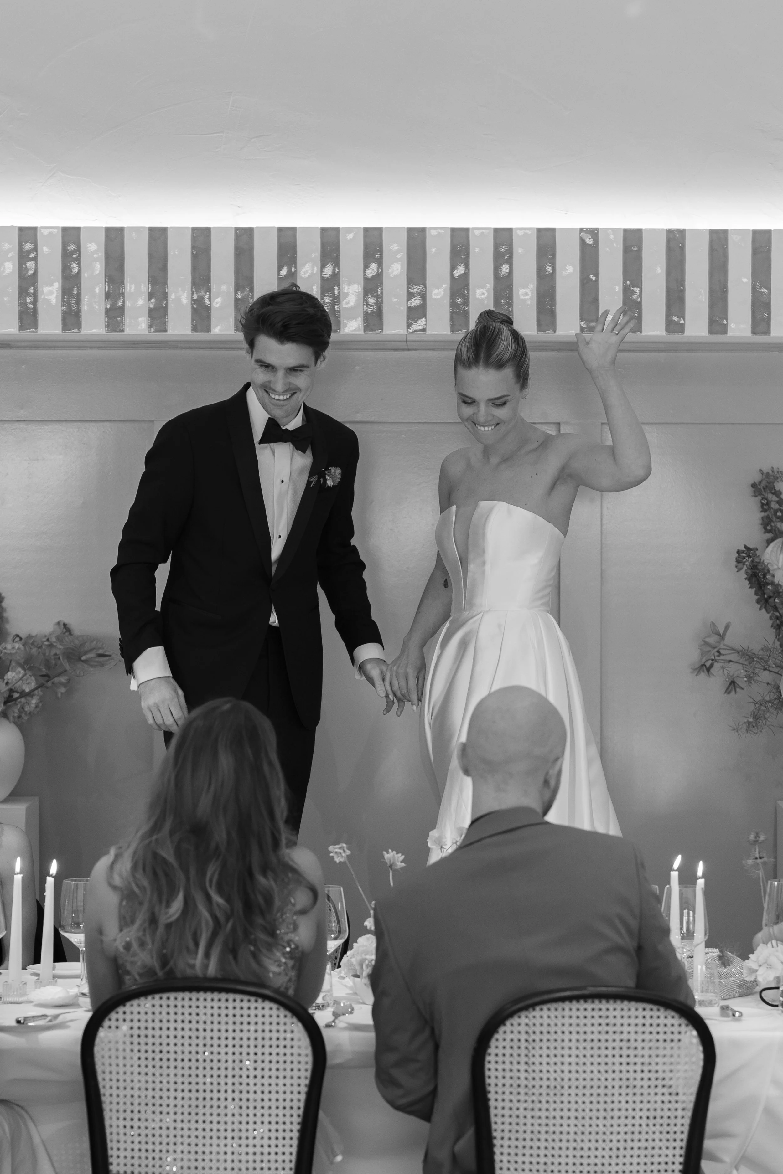 A black and white photo of a bride and groom standing on a table, holding hands and smiling, during their wedding reception. The bride is wearing a strapless wedding dress, and the groom is in a tuxedo. Two guests are seated in front of them, facing 