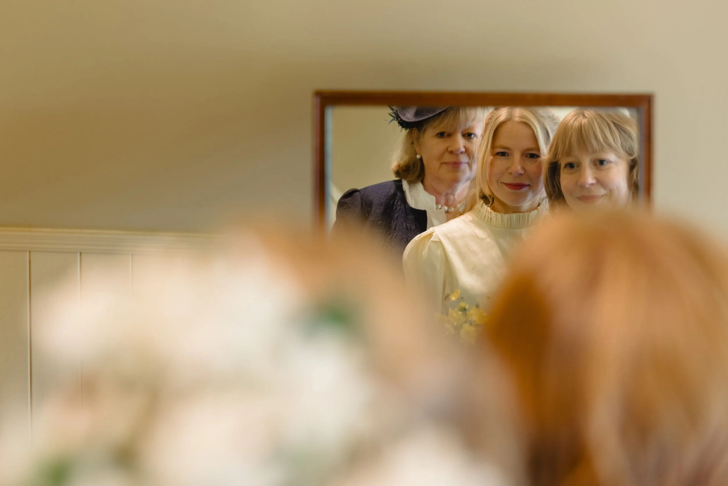 Three women looking into a mirror, with their reflections visible, all smiling.