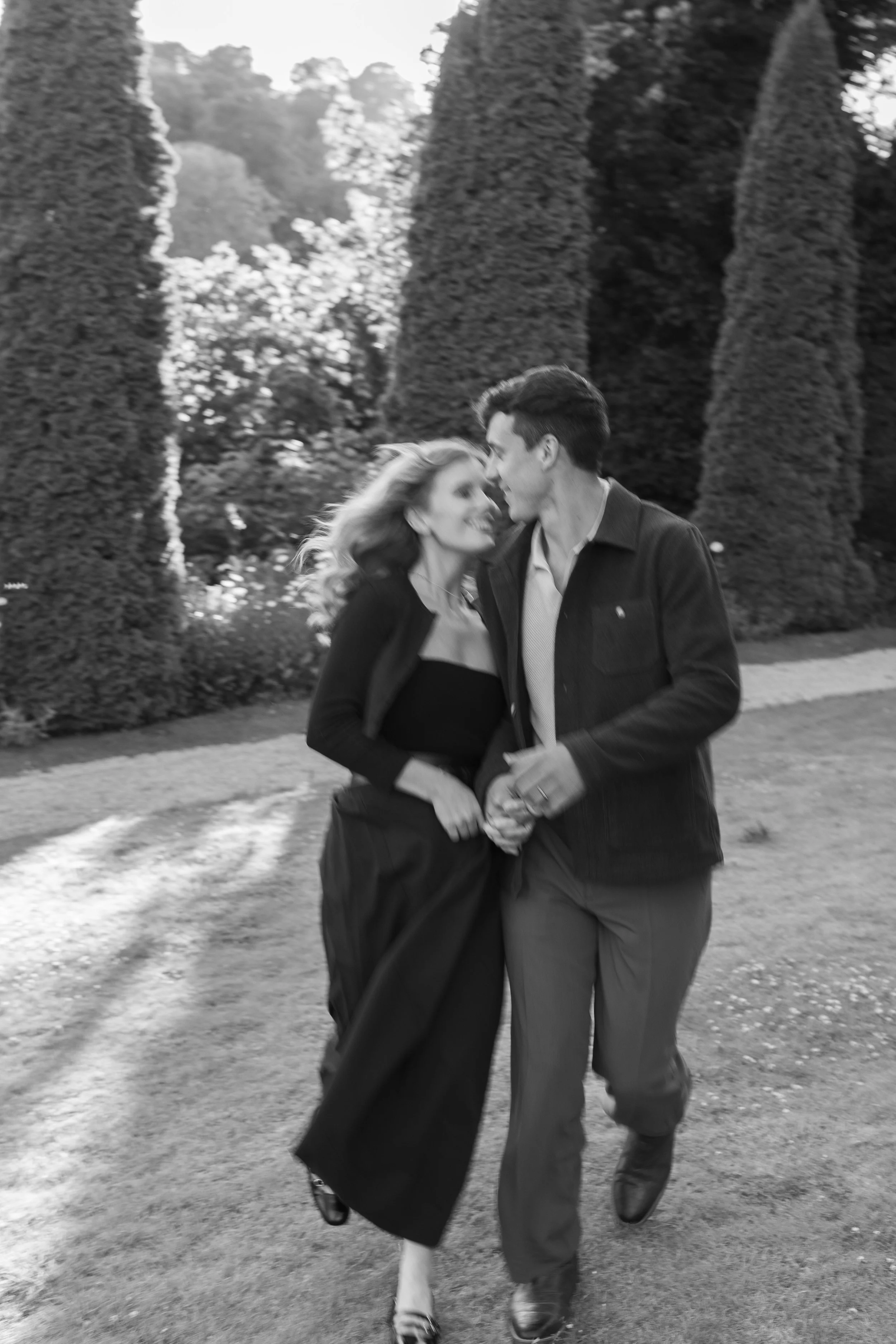 A black and white photo of a couple holding hands and walking outdoors, with tall trees in the background.