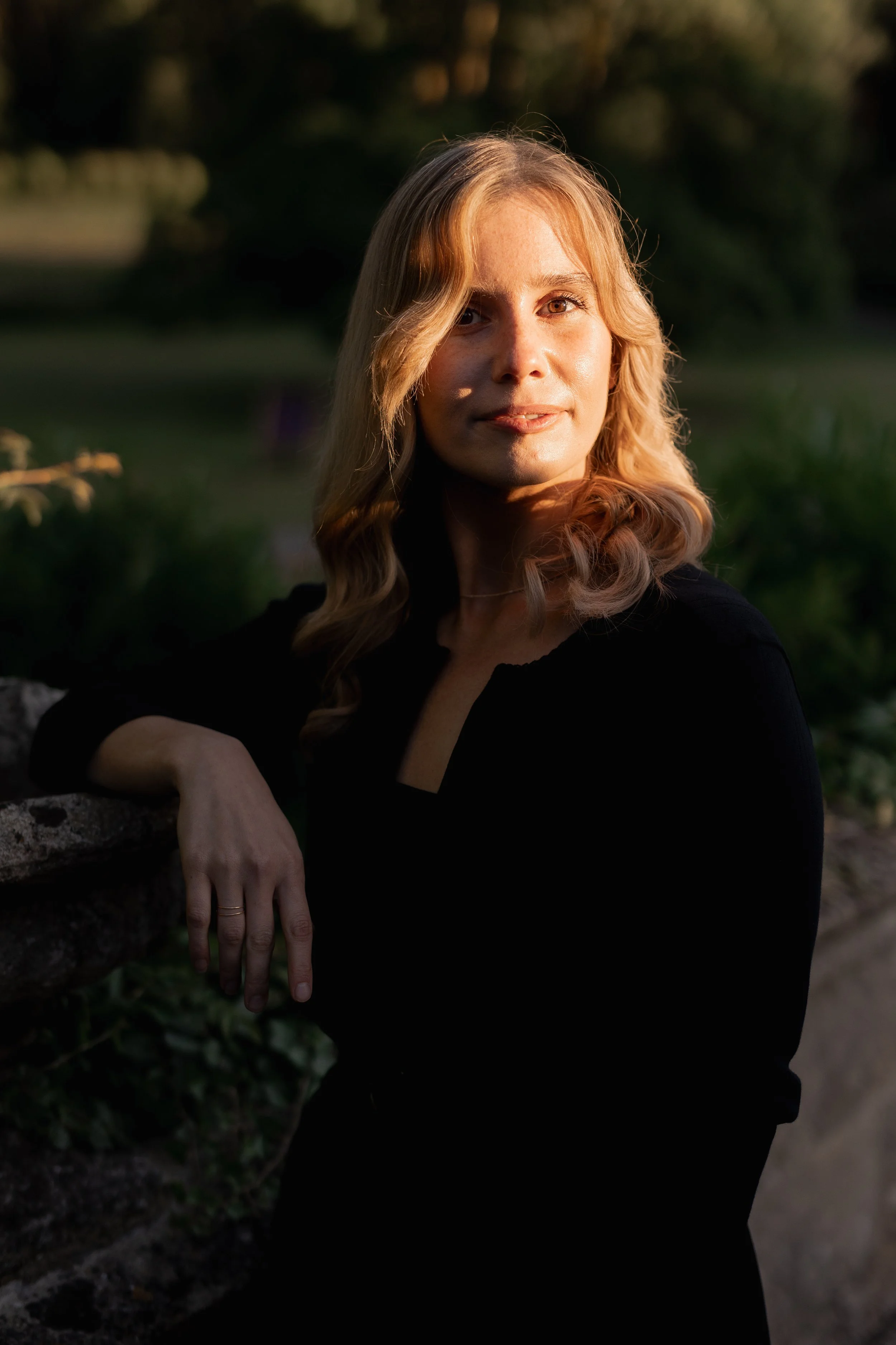 A woman with blonde hair and light skin leaning against a stone wall outdoors during sunset, with soft warm light illuminating her face.