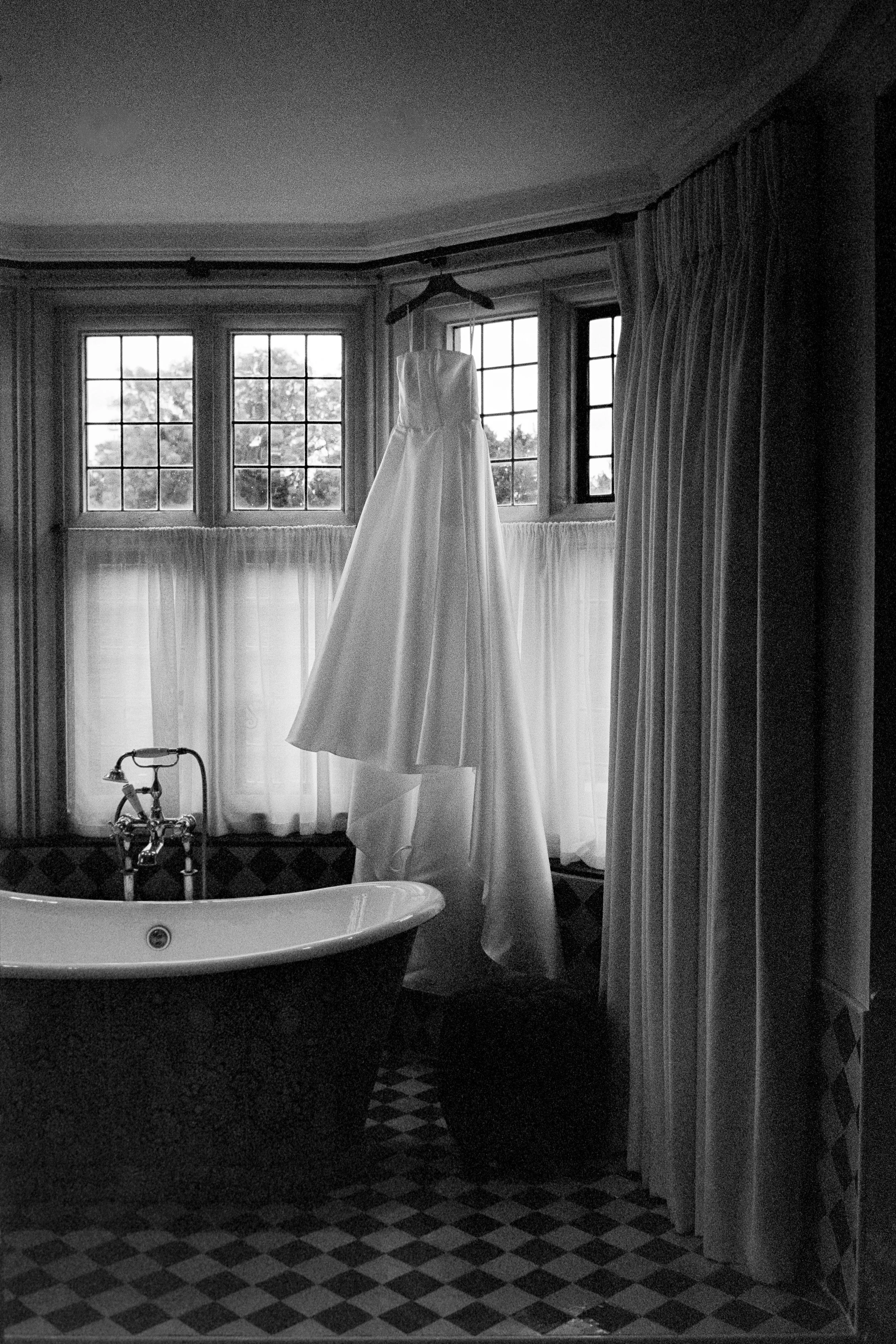 A wedding dress hanging from a hanger in front of a window with a bathtub beneath and curtains on either side.