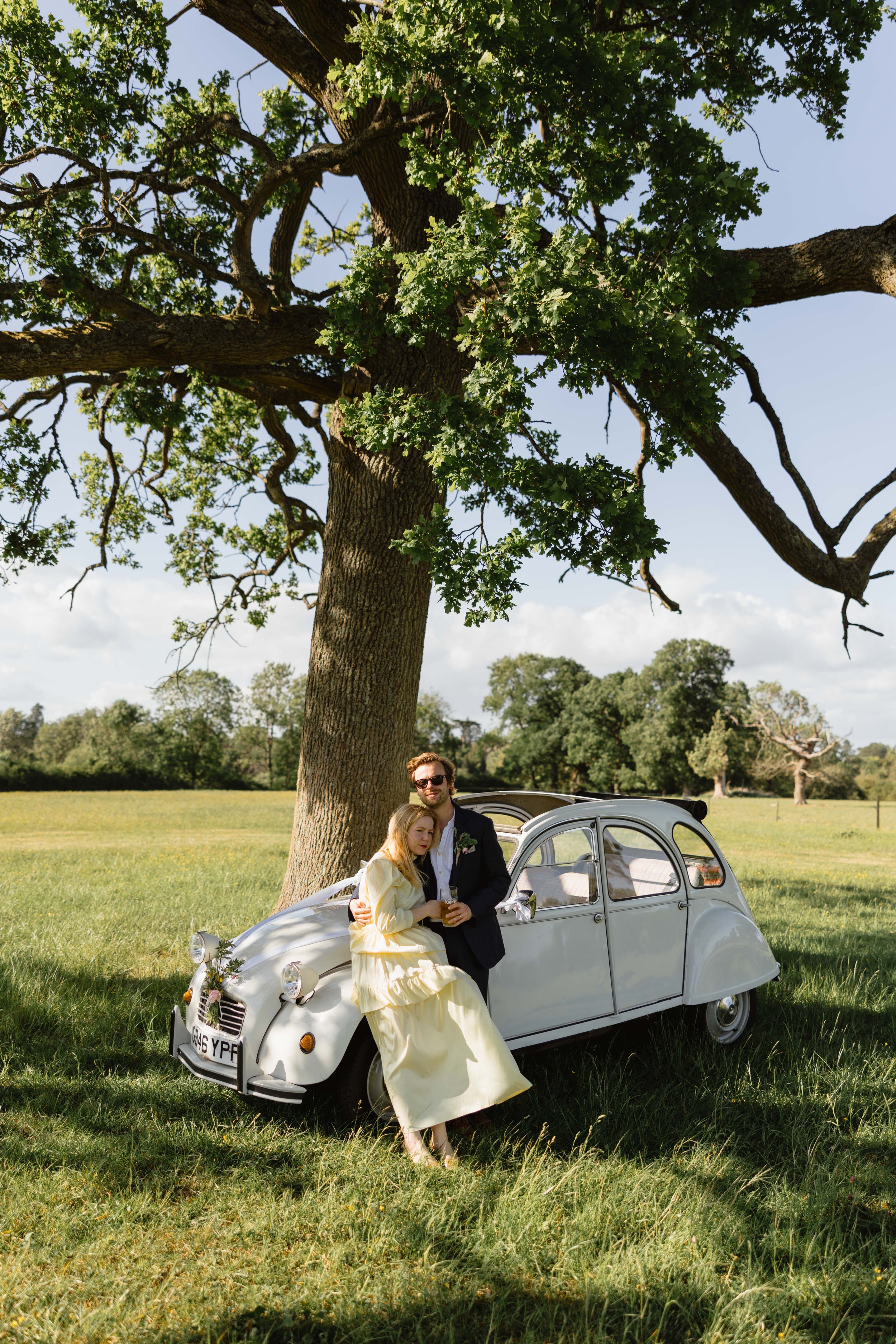 A couple dressed in vintage clothing standing by a classic white Citroën 2CV car under a large tree in a grassy field on a sunny day.