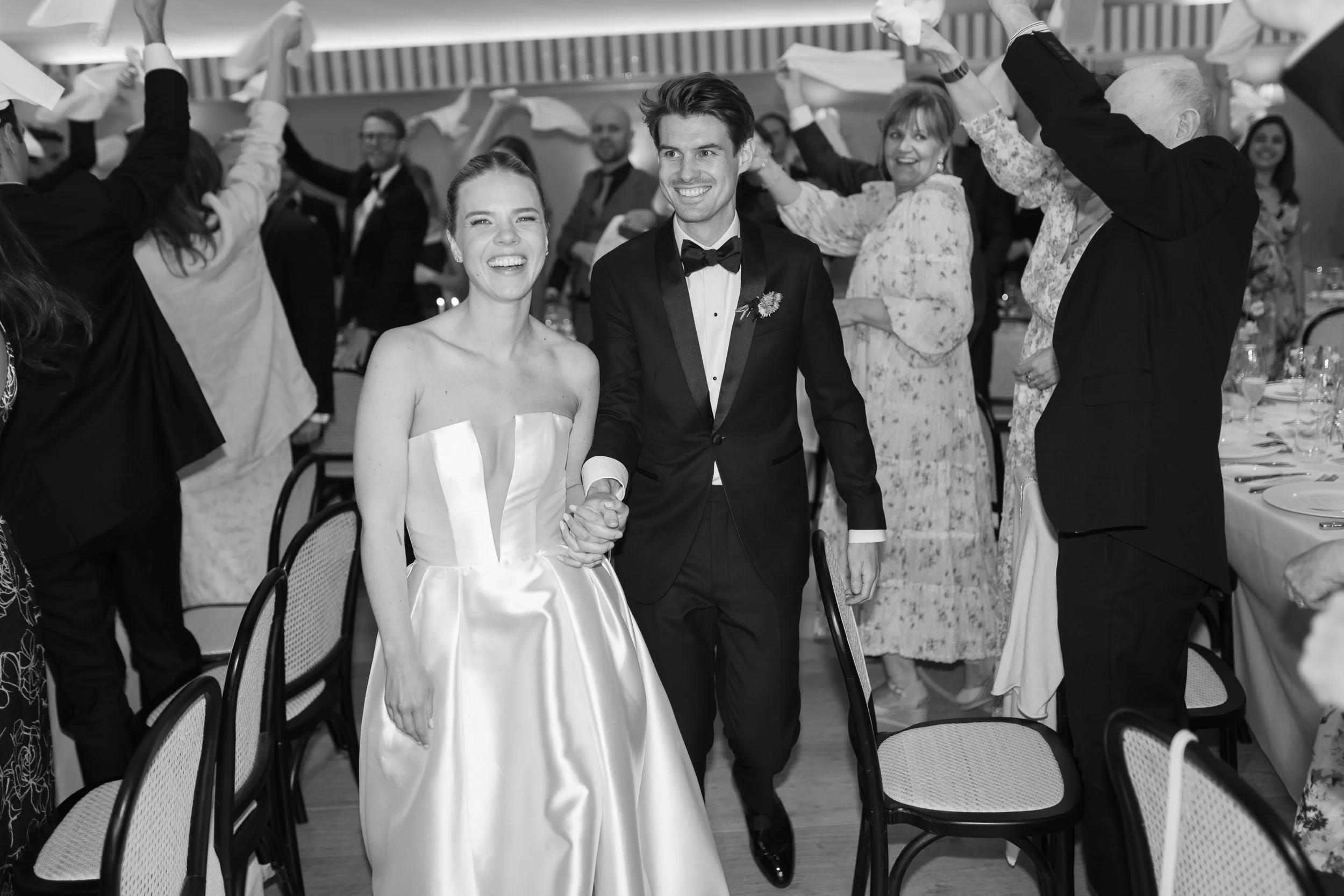 A newlywed couple, a woman in a wedding dress and a man in a tuxedo, walking hand in hand through a celebratory gathering with guests and decorations.