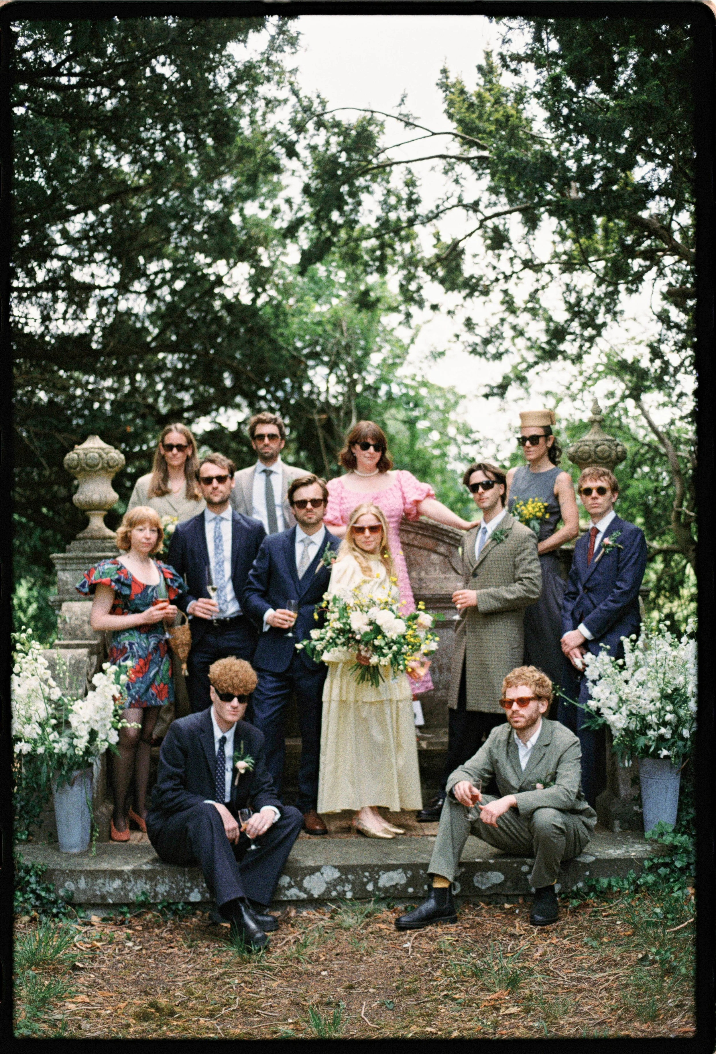 A group of people dressed in vintage clothing posing outdoors in front of stone urns and greenery, for a wedding or special event.