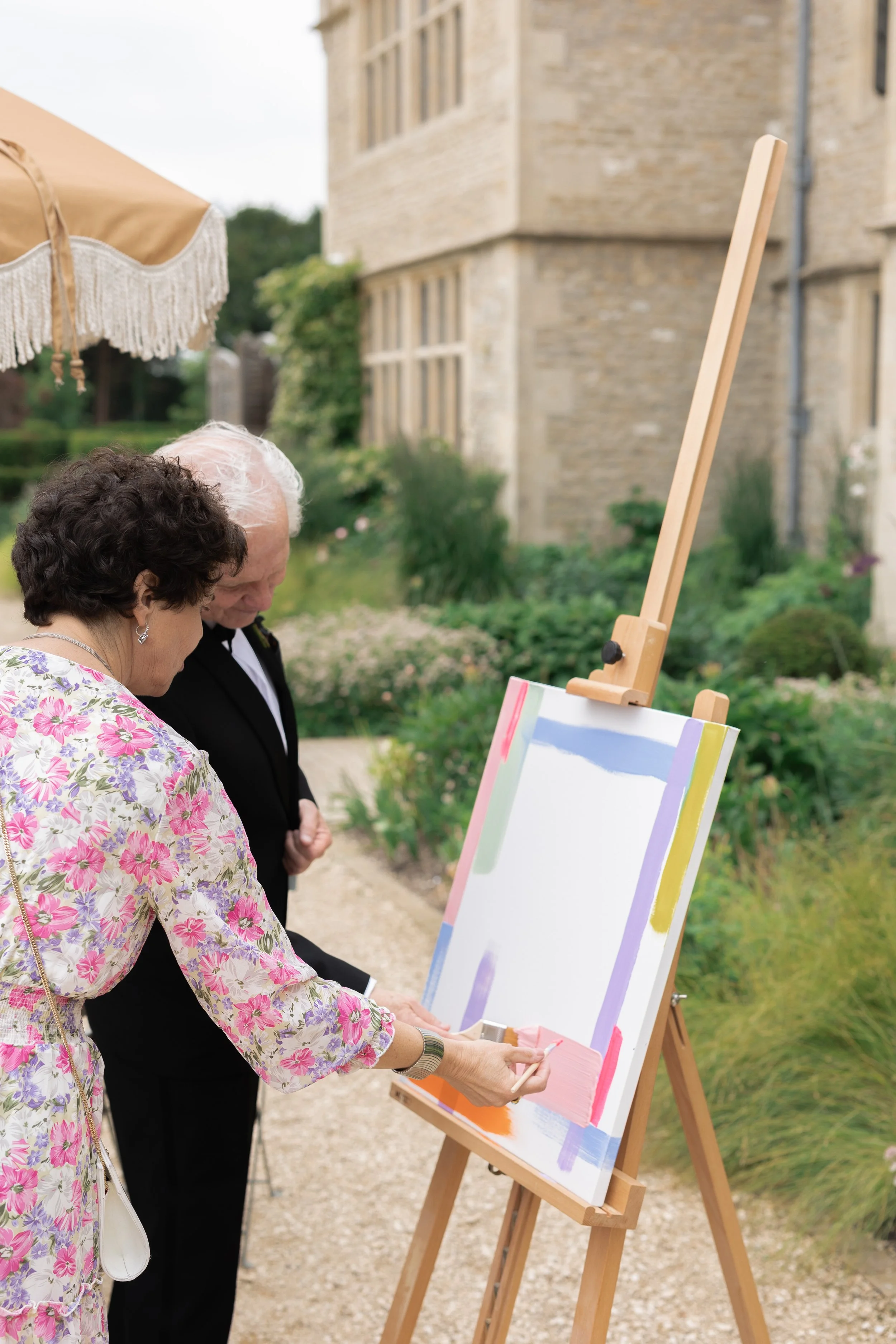 Two people, a man and a woman, are painting on a large canvas on an easel outdoors. The woman is wearing a floral dress and the man is in a tuxedo. They are standing in a garden area near a building.