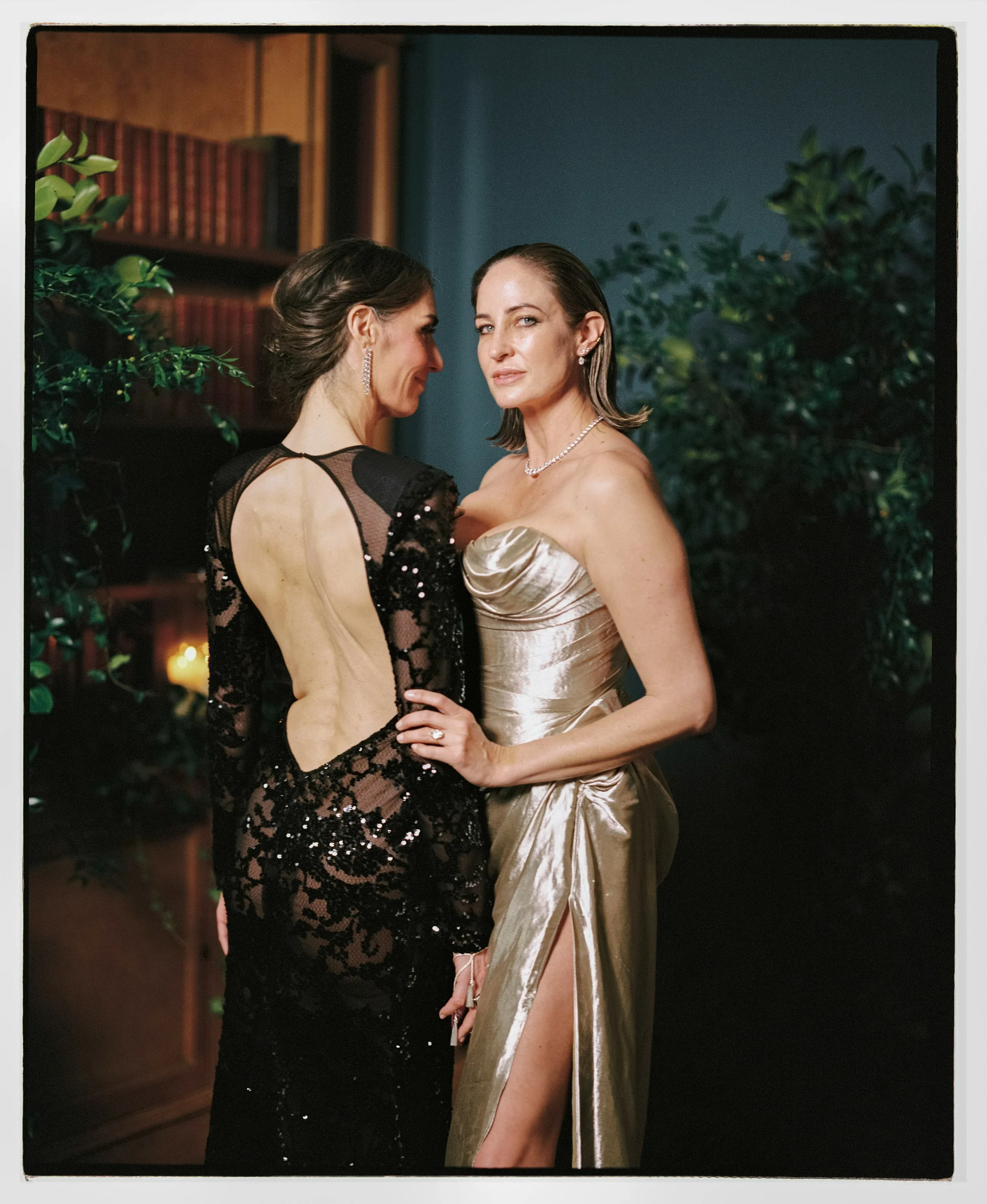 The brides in formal evening gowns standing close together, with one woman in a black, lace, backless dress and the other in a shiny, strapless, high-slit gown. They are indoors, surrounded by green plants and dark wood bookshelves.