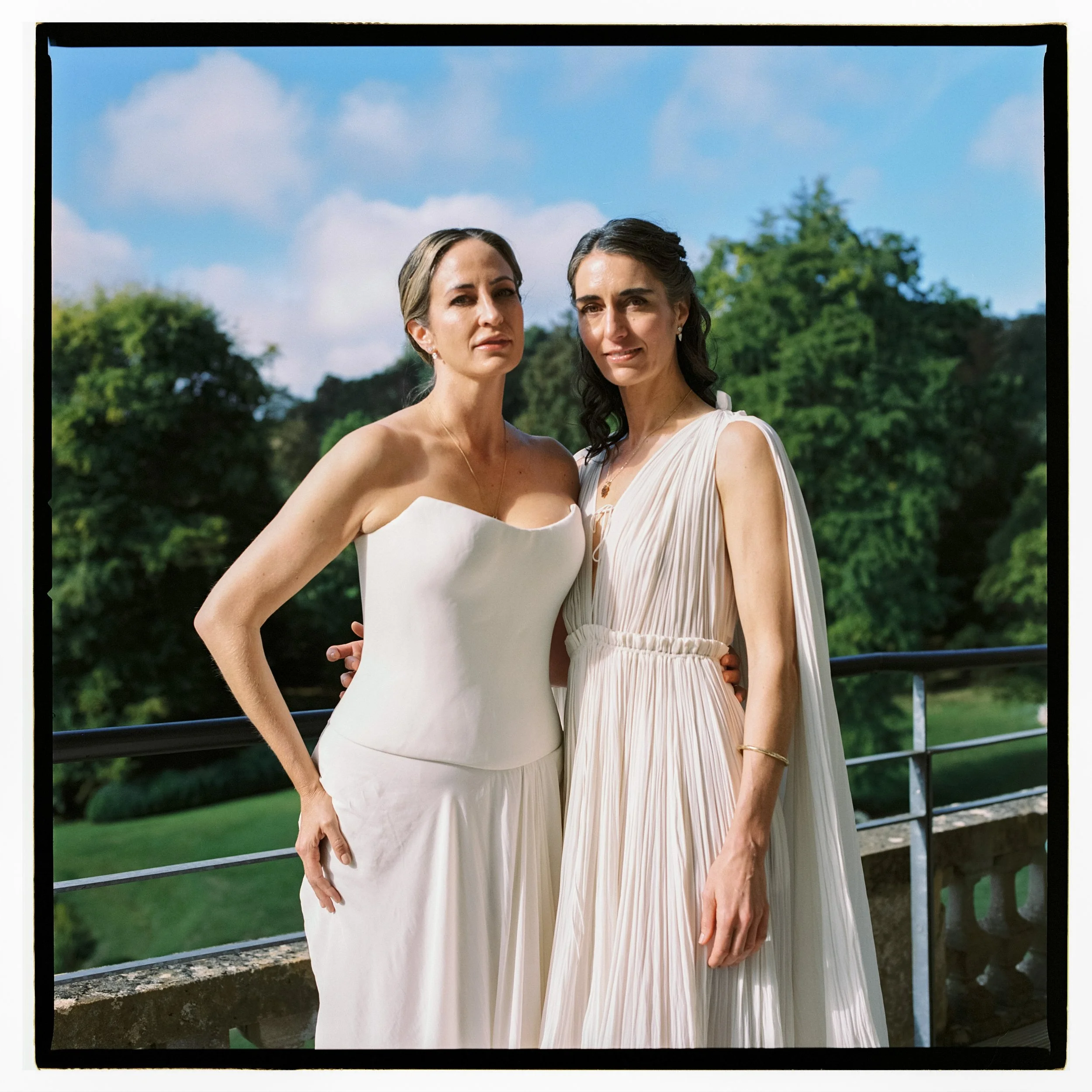 Two women in wedding dresses standing outdoors on a balcony with greenery and trees in the background.