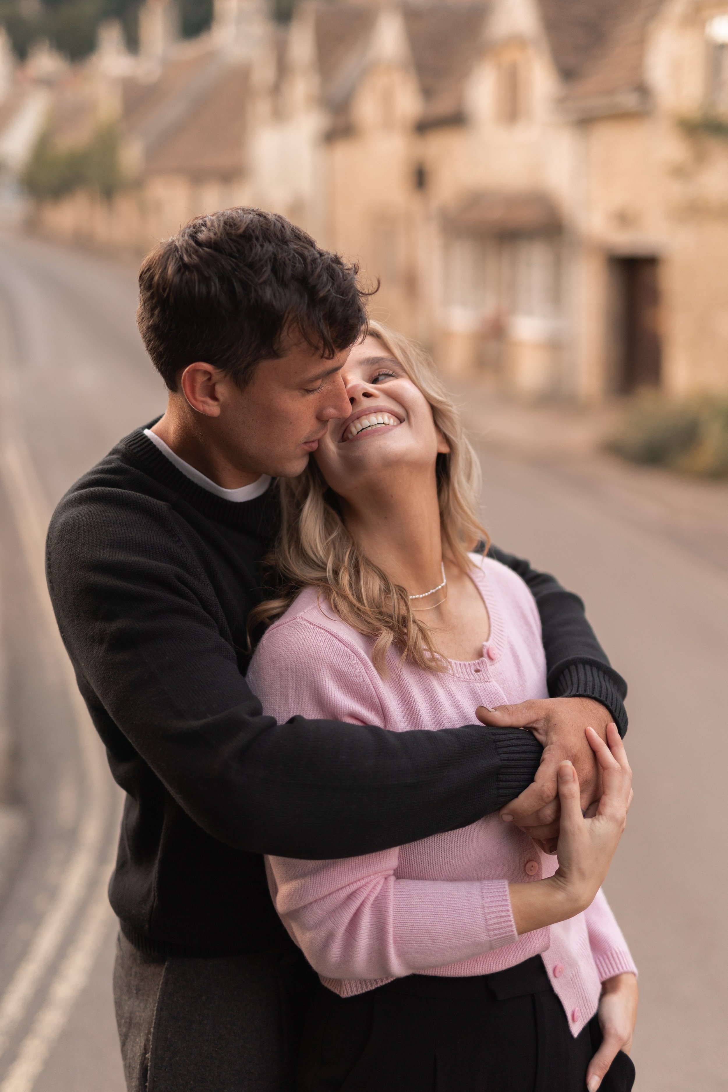 A young man and woman embrace warmly on a quiet street with old buildings in the background. The man leans in to kiss the woman's cheek as she smiles happily.
