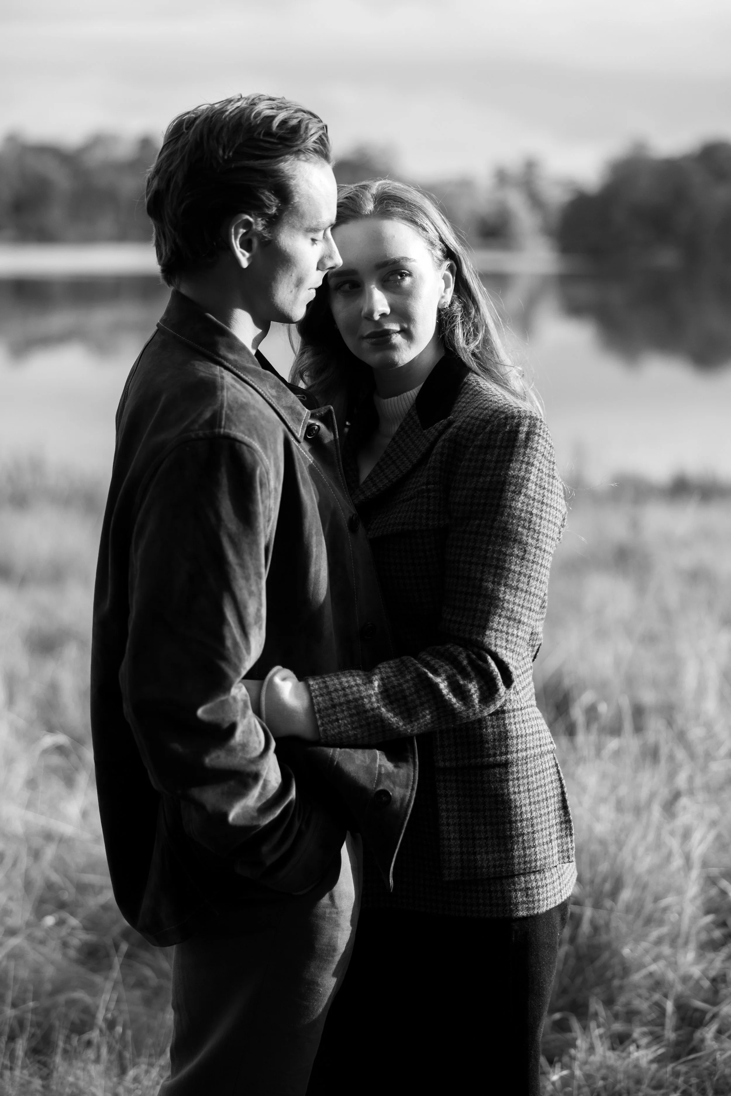 Black and white photo of a man and woman standing close by a lake, with the man in a leather jacket and the woman in a checkered blazer, facing each other outdoors.
