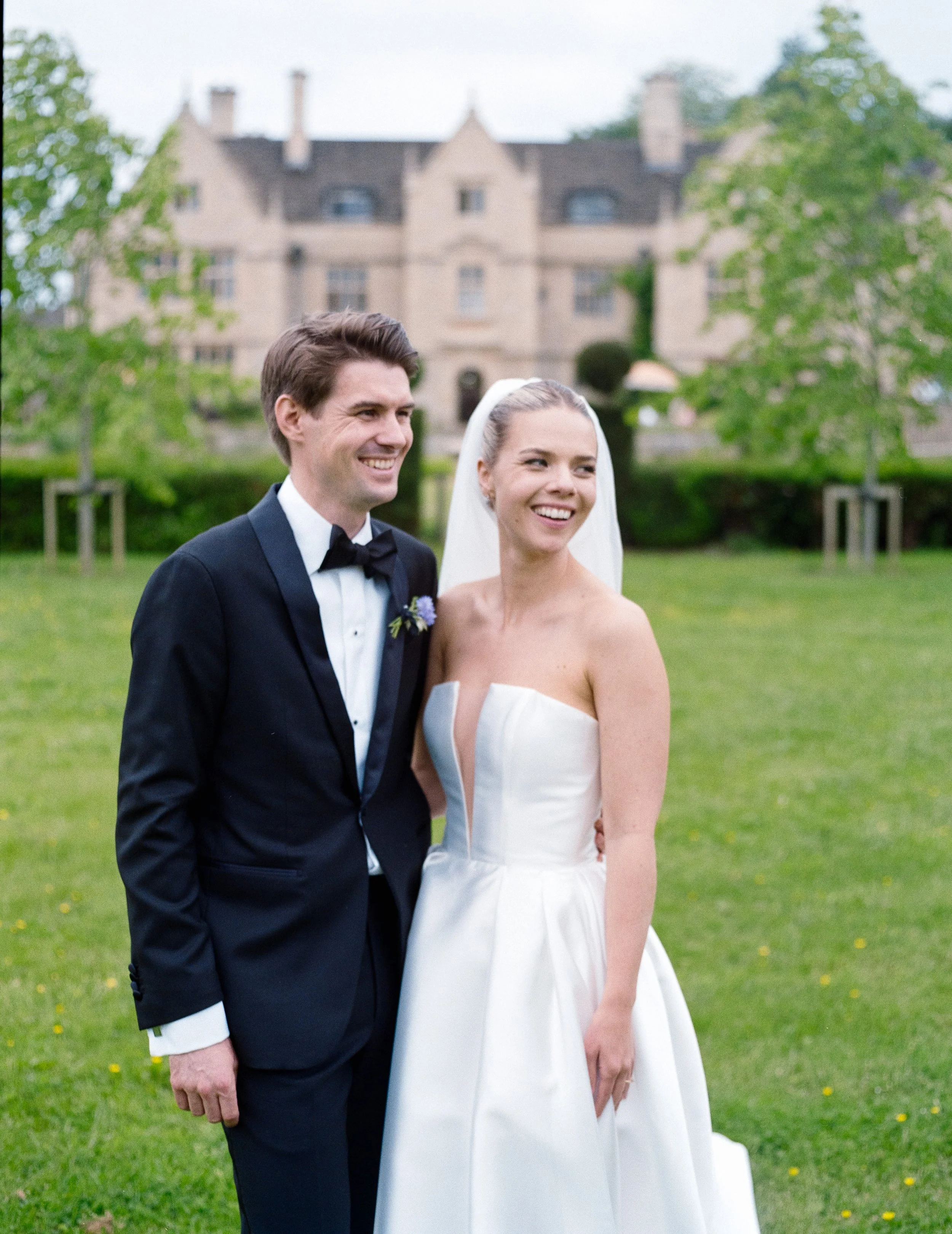 A bride and groom standing outside on green grass, smiling, with a large historic building in the background. She is wearing a strapless white wedding gown with a veil, and he is in a black tuxedo with a bow tie.