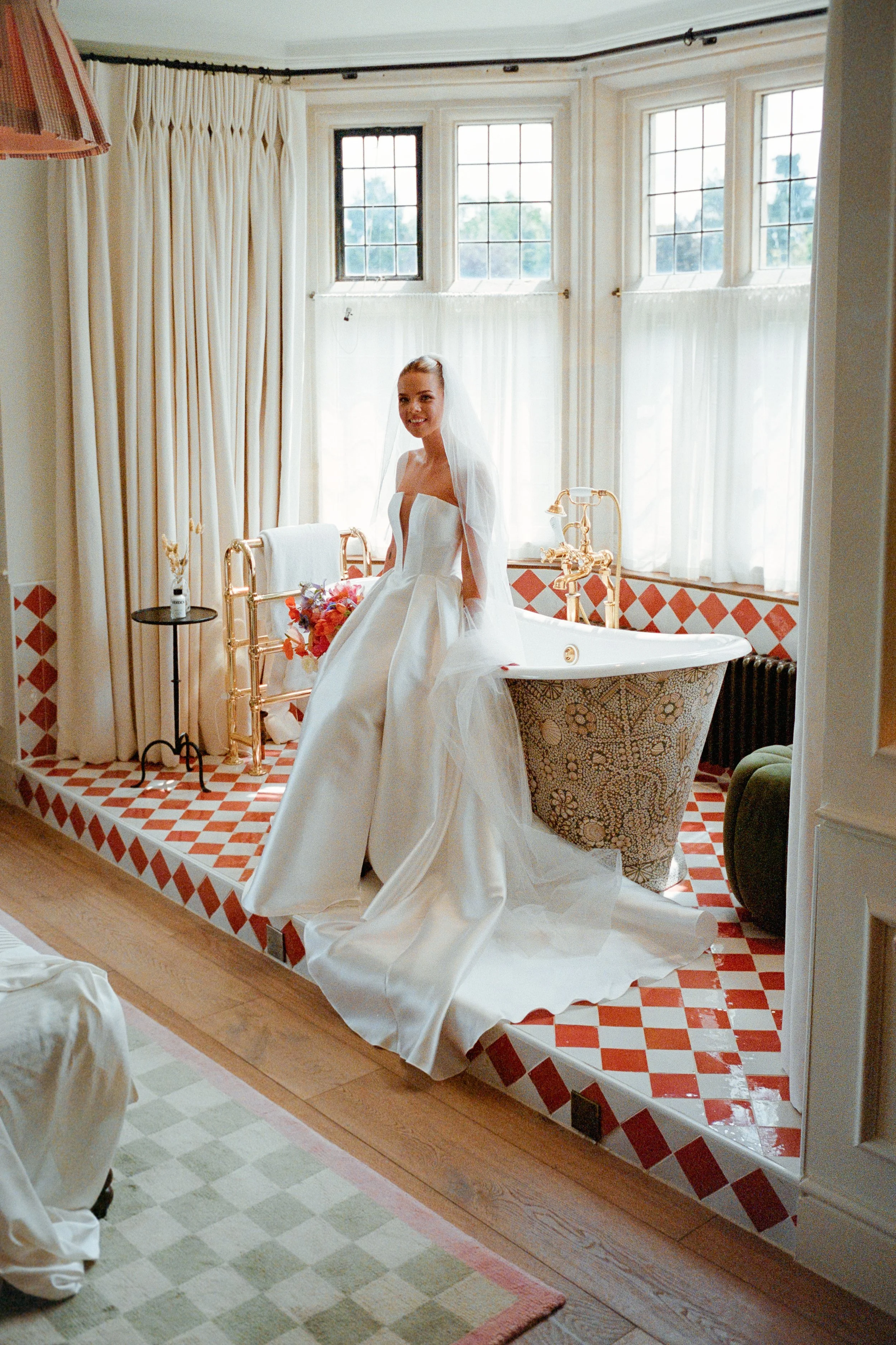 A bride in a white wedding gown sitting on the edge of a clawfoot bathtub in a bright, vintage-style bathroom with patterned tiles, large windows, and gold fixtures.
