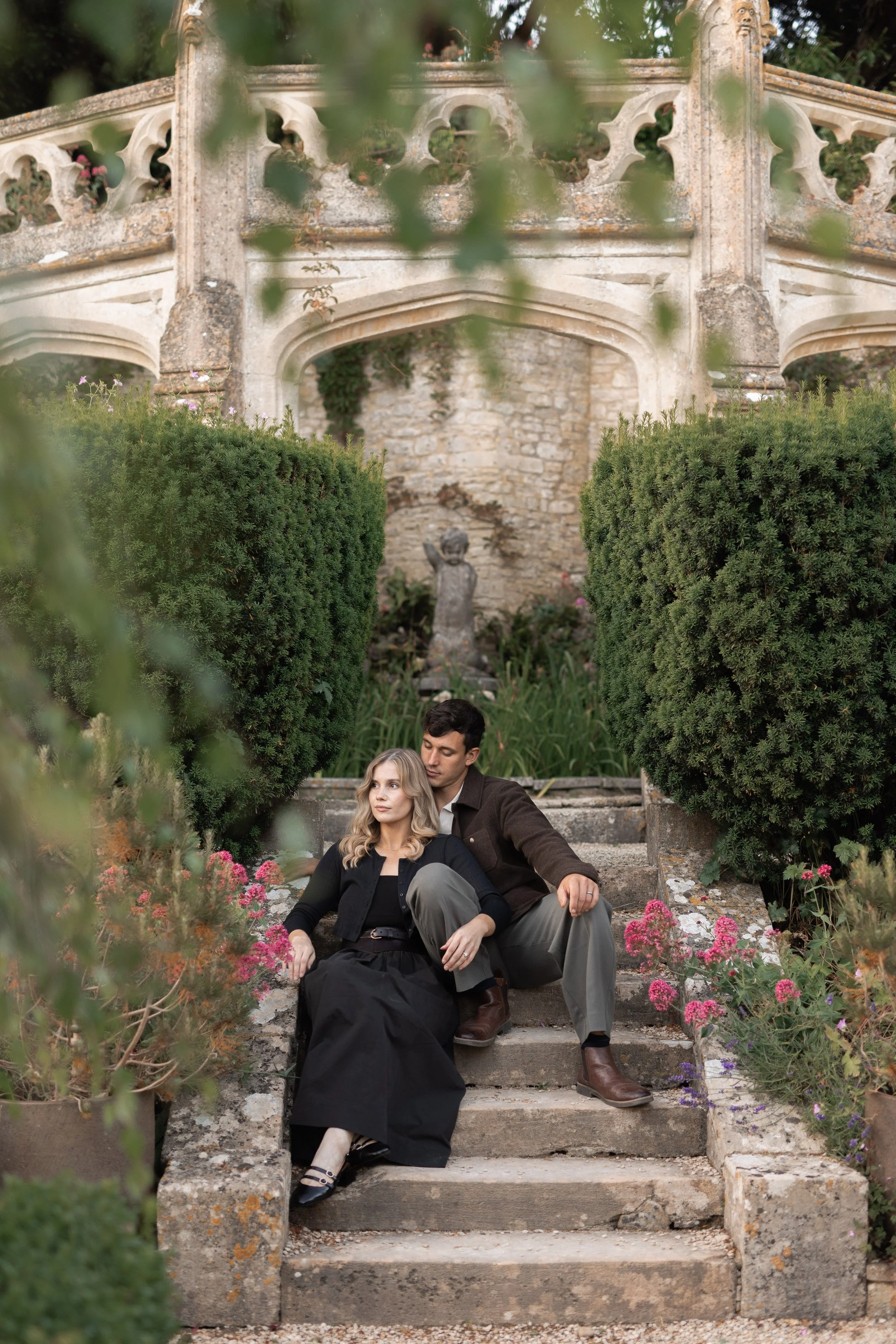 A man and a woman sitting on stone steps in a garden with green bushes, flowers, and an old stone wall with a sculpture behind them.