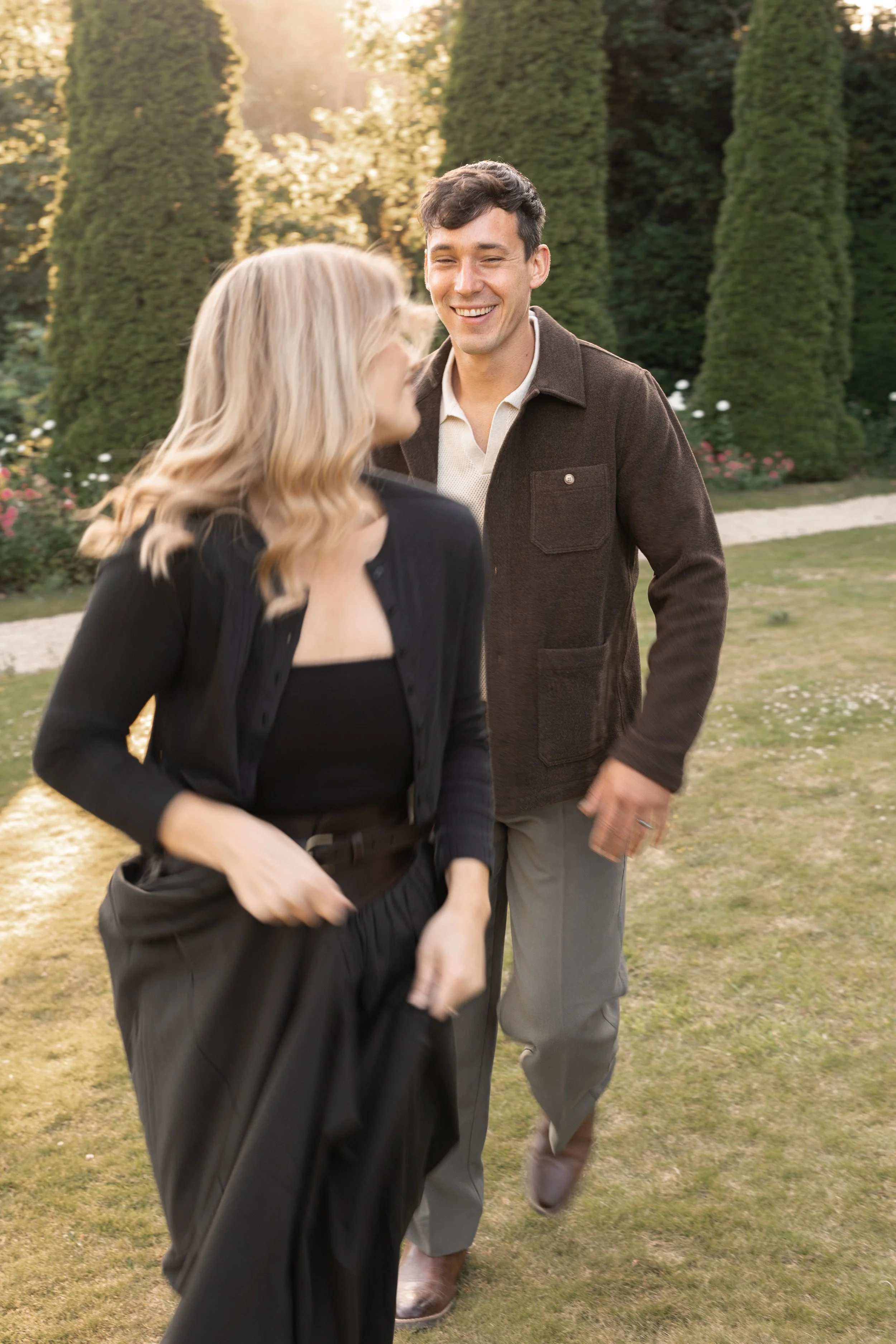 A man and woman walking and smiling outdoors in a garden with tall trees and flowers in the background.