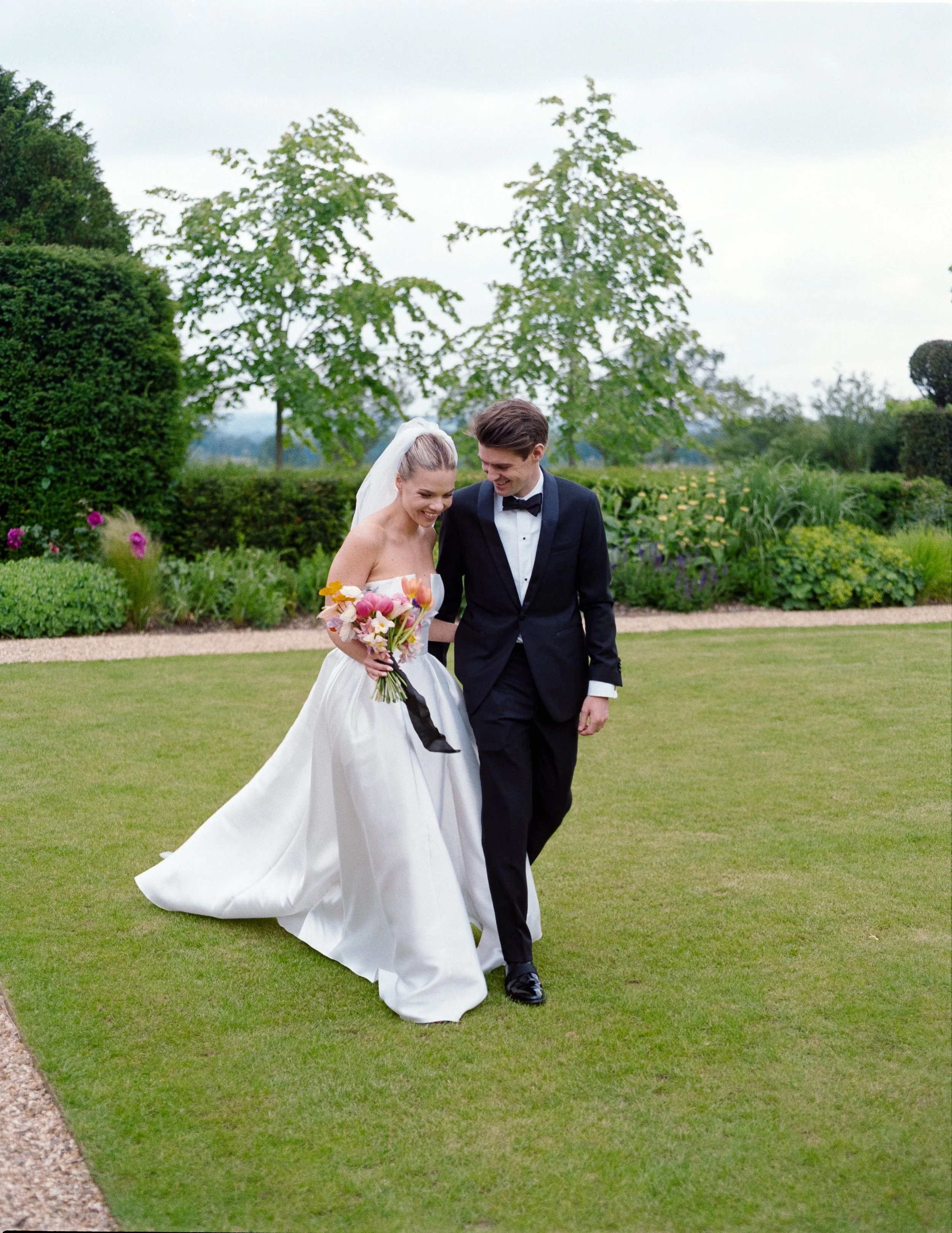 A bride and groom walking on a lawn during a wedding, smiling and holding hands, with trees and bushes in the background.
