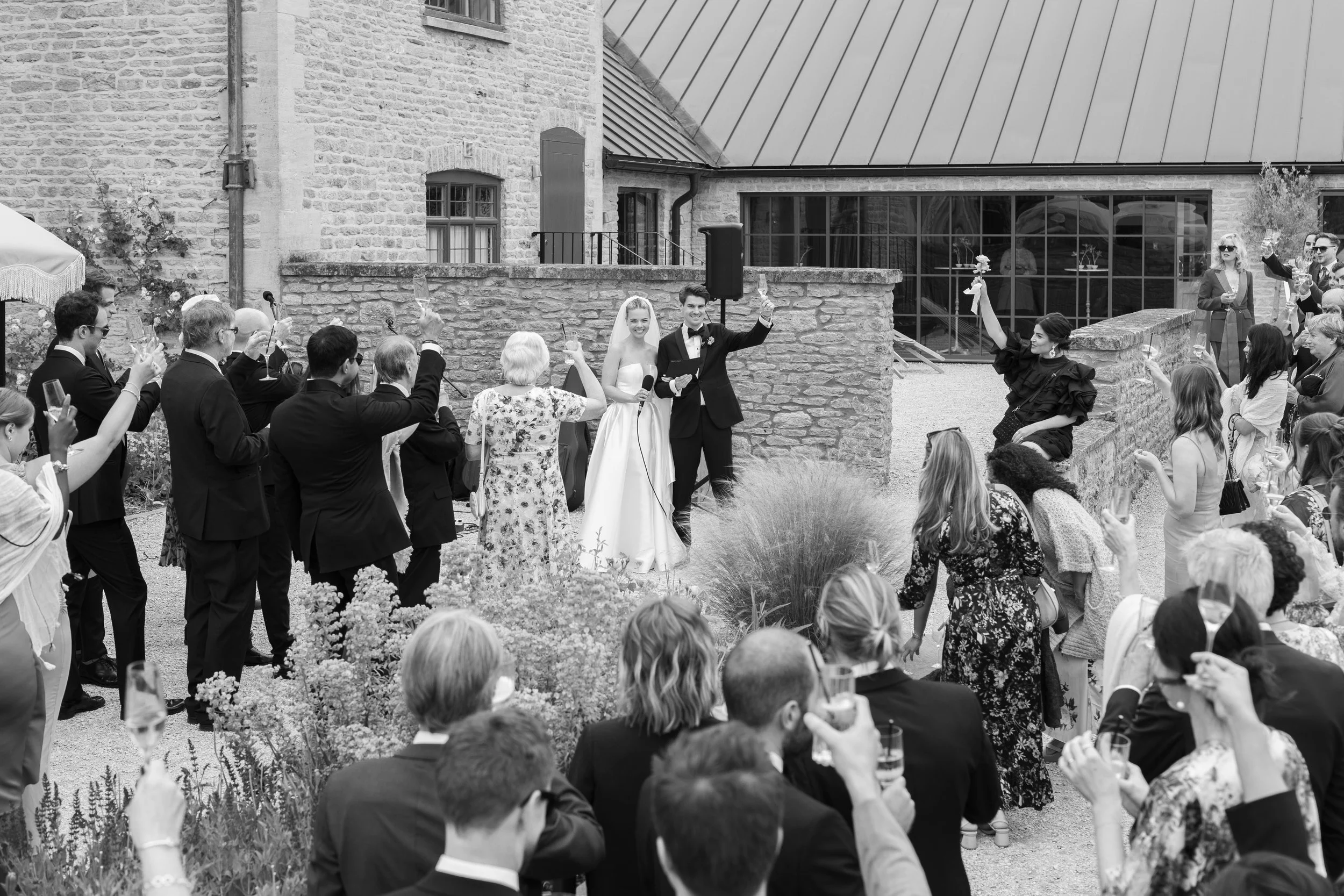 A wedding celebration outdoors with a bride and groom at the center, surrounded by guests raising glasses for a toast, in front of a stone building.