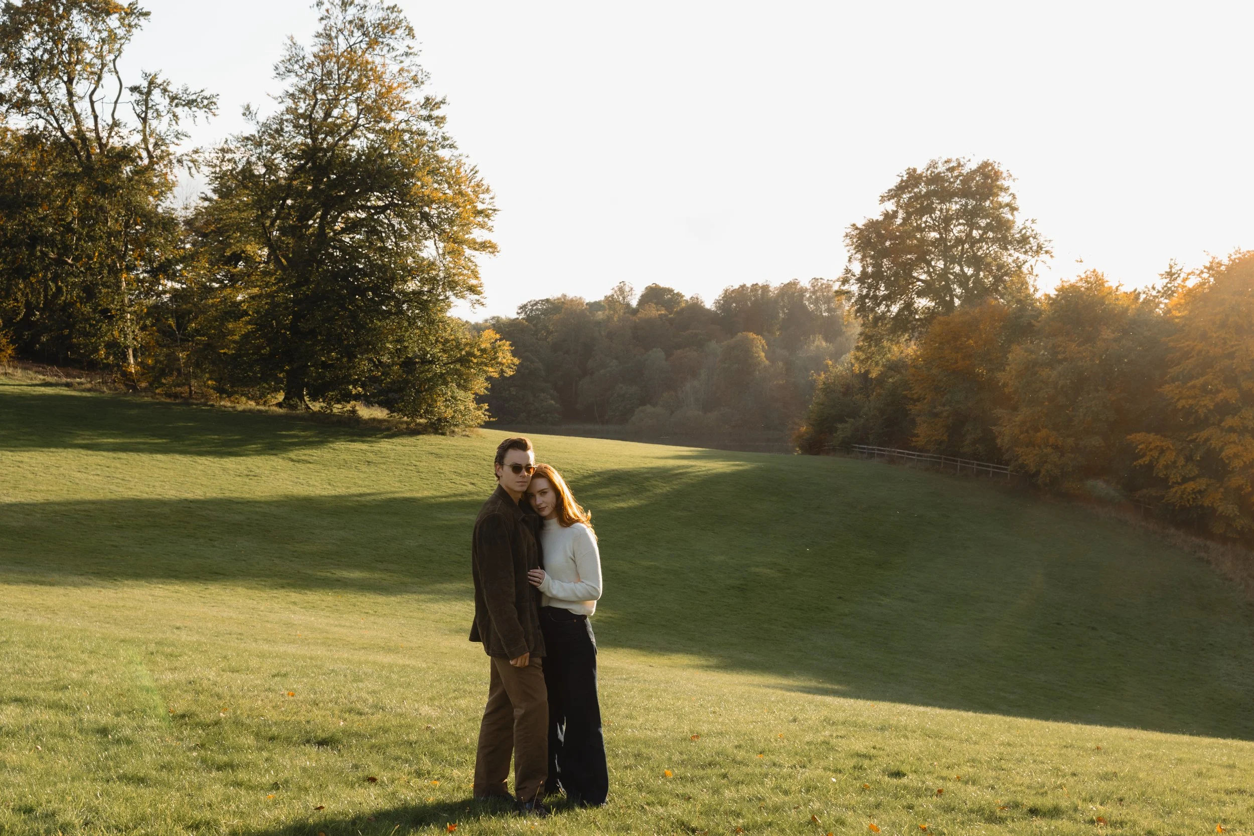 A couple standing on a grassy hillside in a park, embracing, with trees in the background and sunlight coming from the right.