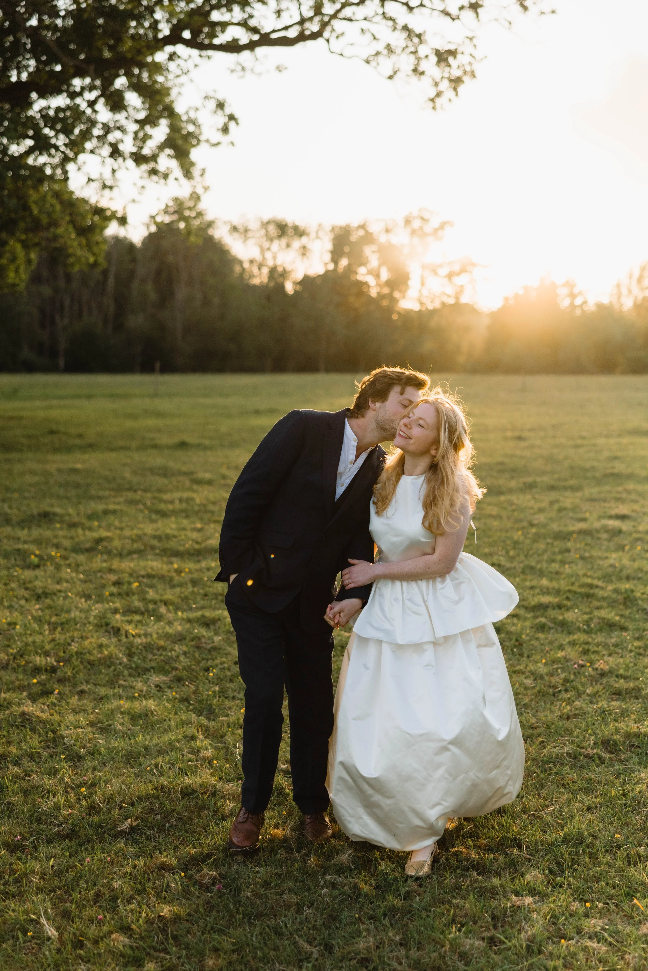 A couple dressed in formal wedding attire standing in a grassy field at sunset, sharing a tender moment with the man kissing the woman on the cheek, both smiling.