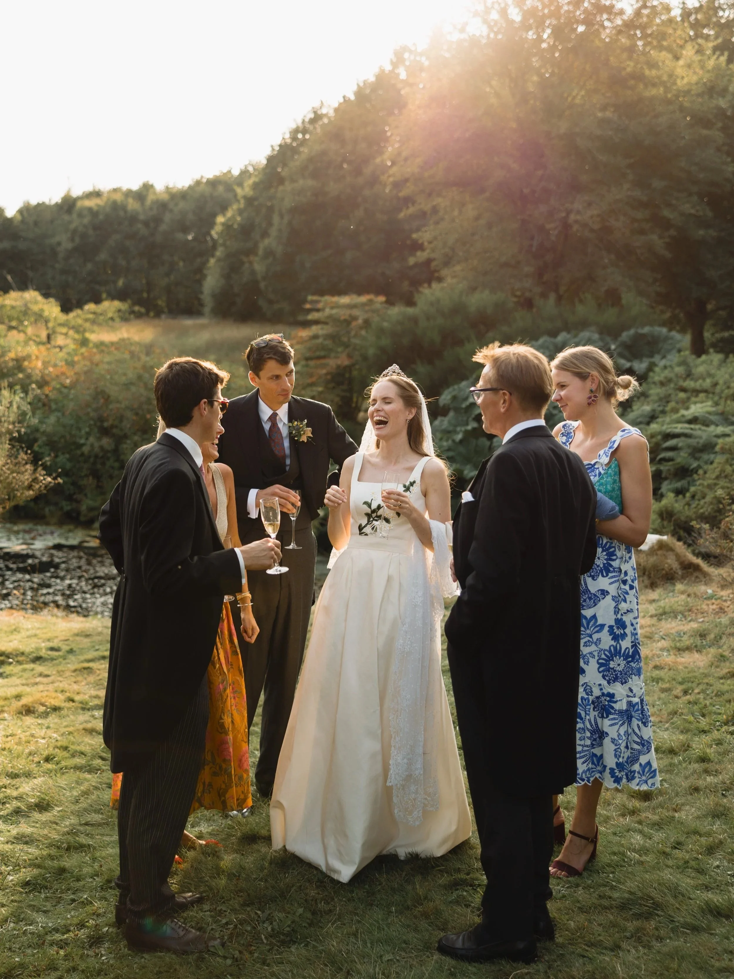 Group of people celebrating outdoors in formal attire, likely at a wedding, with a bride in a white gown and tiara, holding a champagne glass, surrounded by friends and family, with lush greenery and trees in the background during sunset.