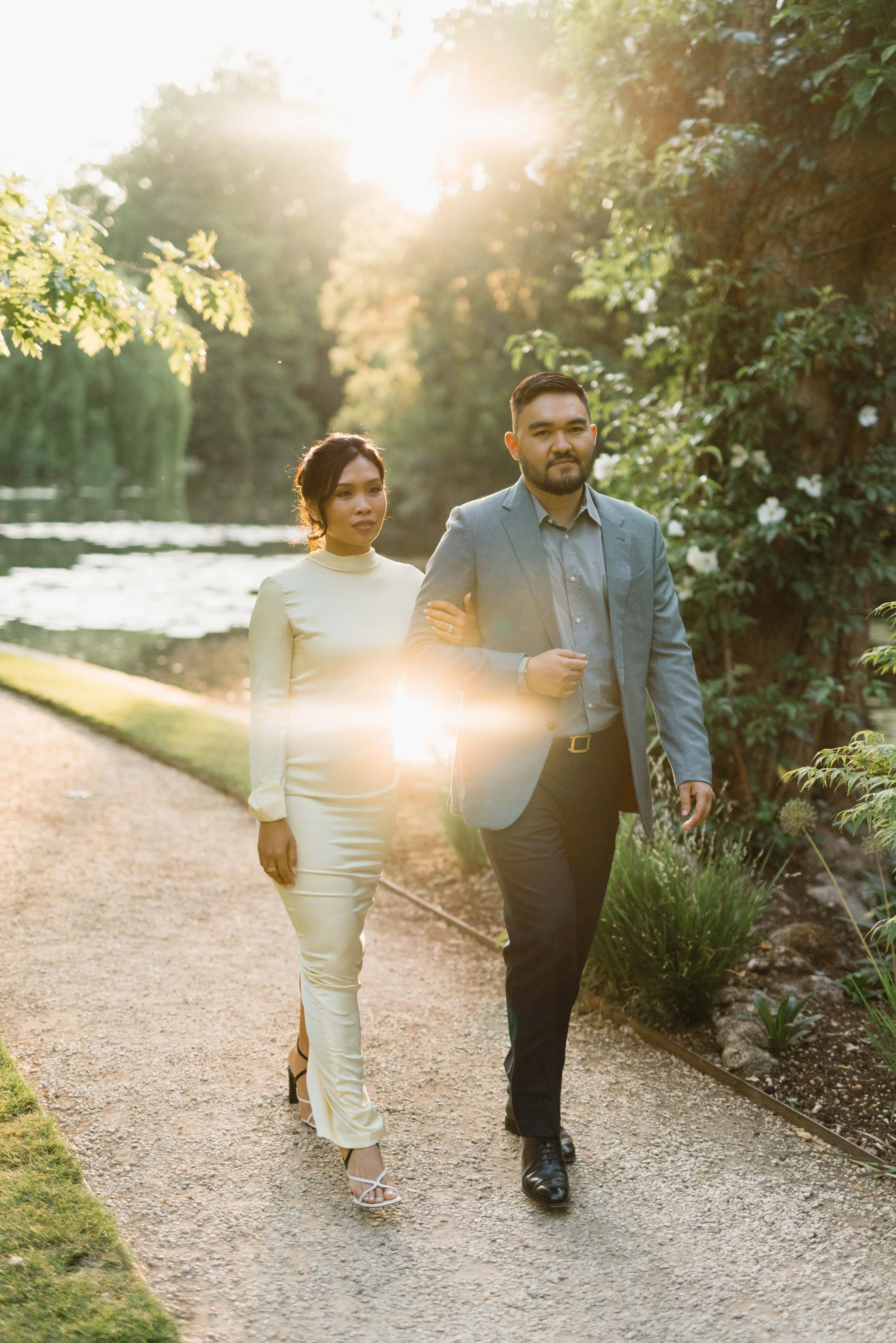A man and woman walking together on a gravel path beside a pond or lake, surrounded by lush greenery, with sunlight filtering through the trees, creating a warm, serene atmosphere.