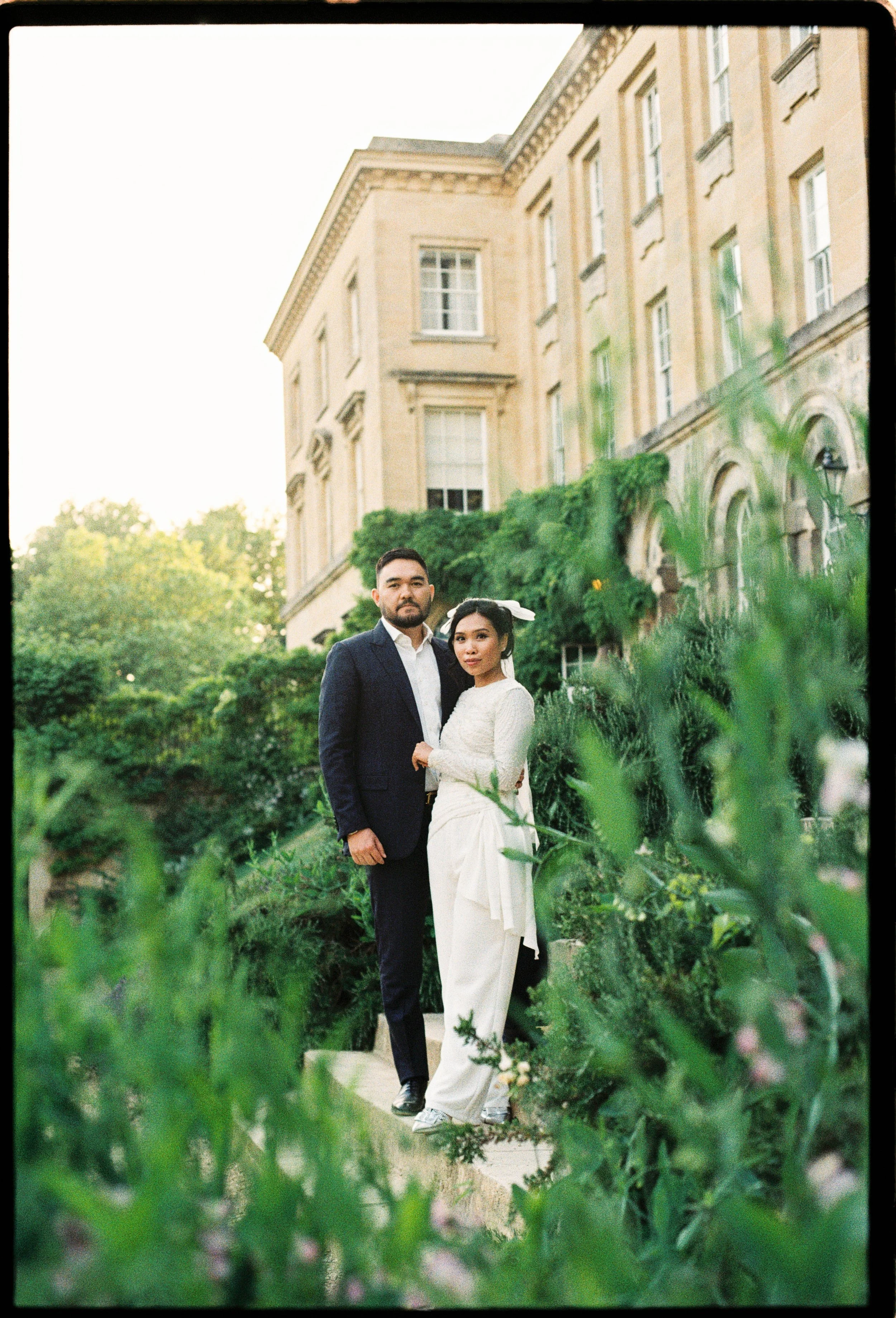 A couple standing on a garden path in front of a historic building, surrounded by greenery, at sunset.