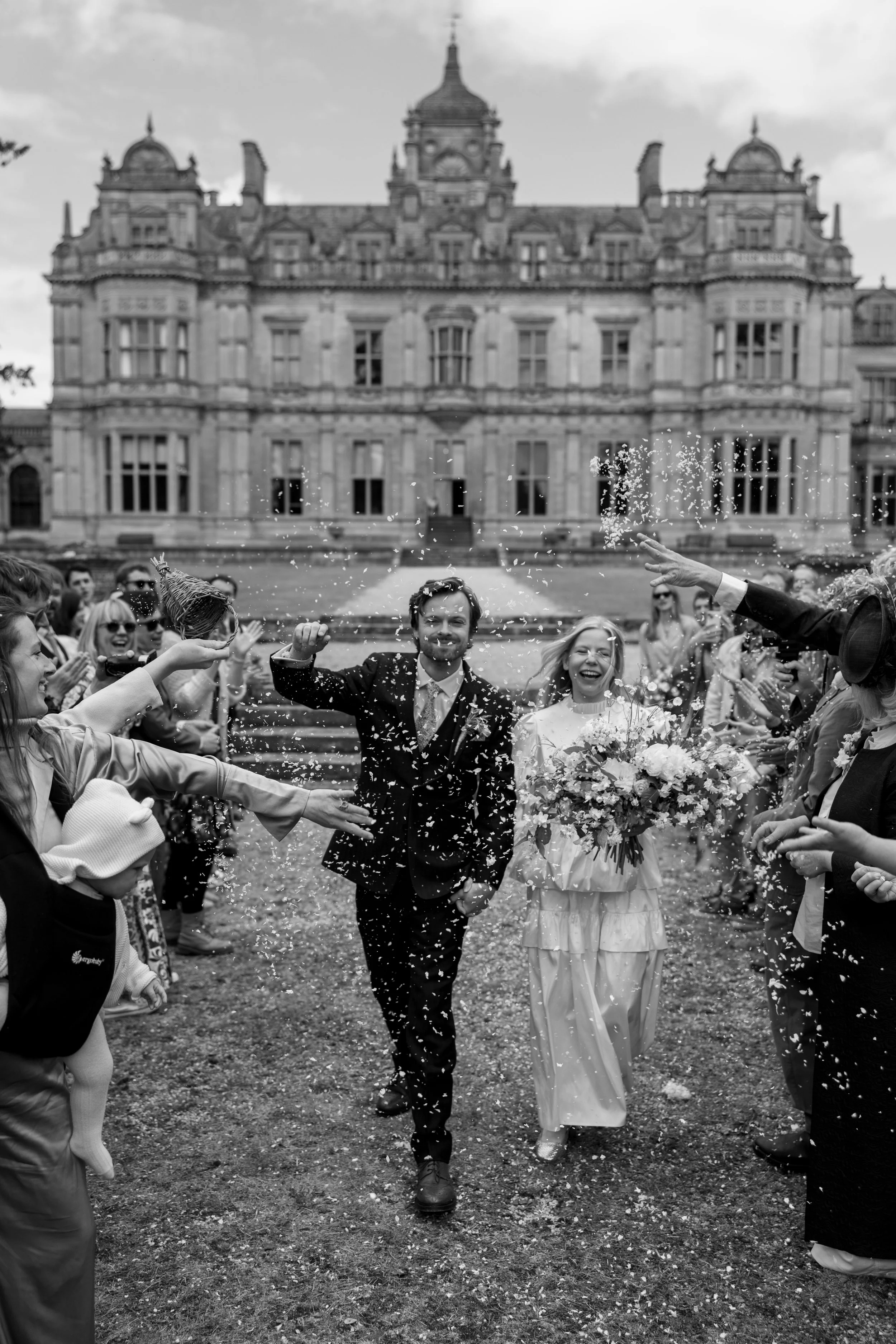 The photo shows a newly married couple walking through a crowd of people celebrating with confetti and laughter outside a large historic building.