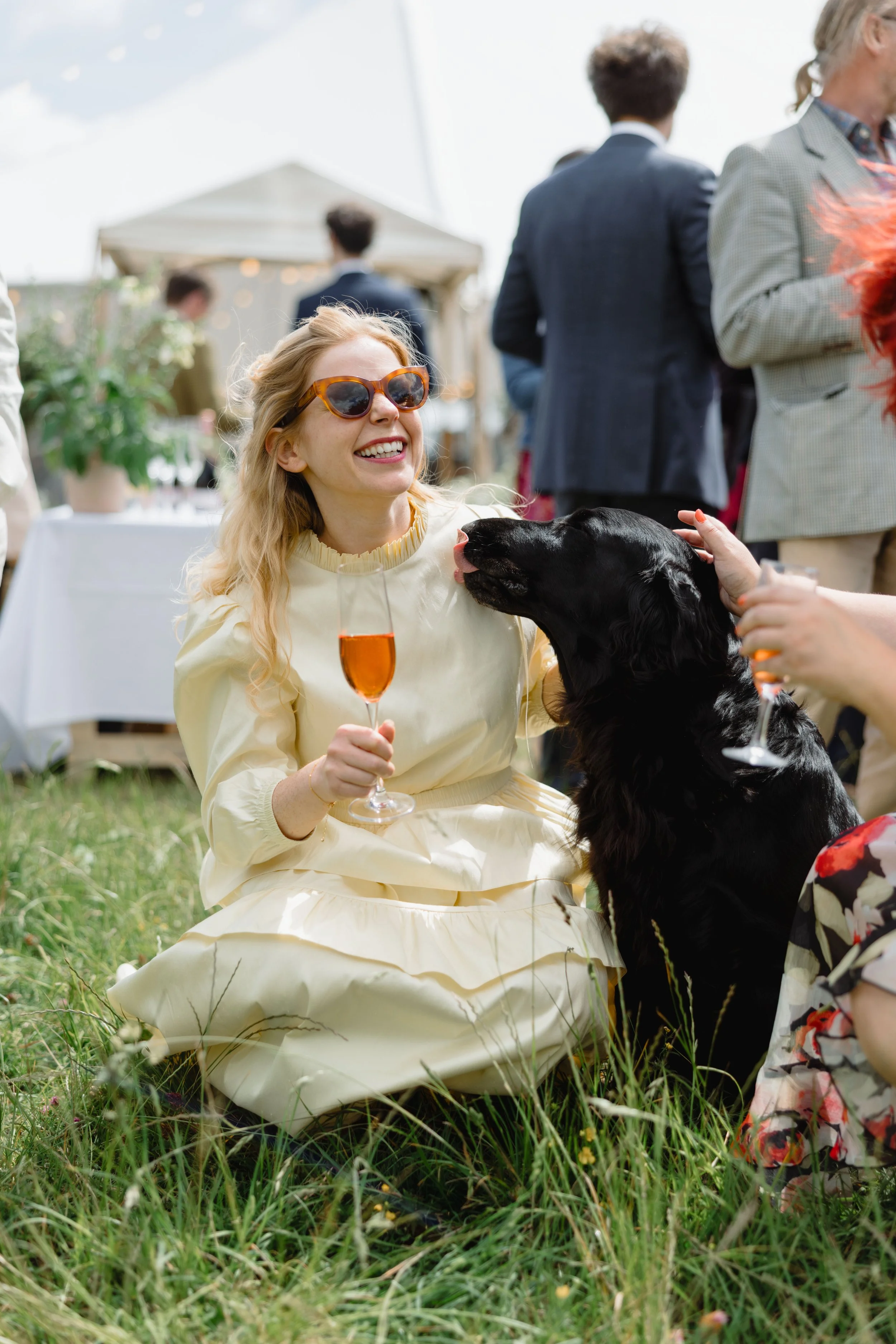 A woman with blonde hair, wearing sunglasses and a cream-colored dress, sitting on grass at an outdoor social gathering, smiling while a black dog licks her face, holding a glass of orange-colored drink, with other people in the background and a tent