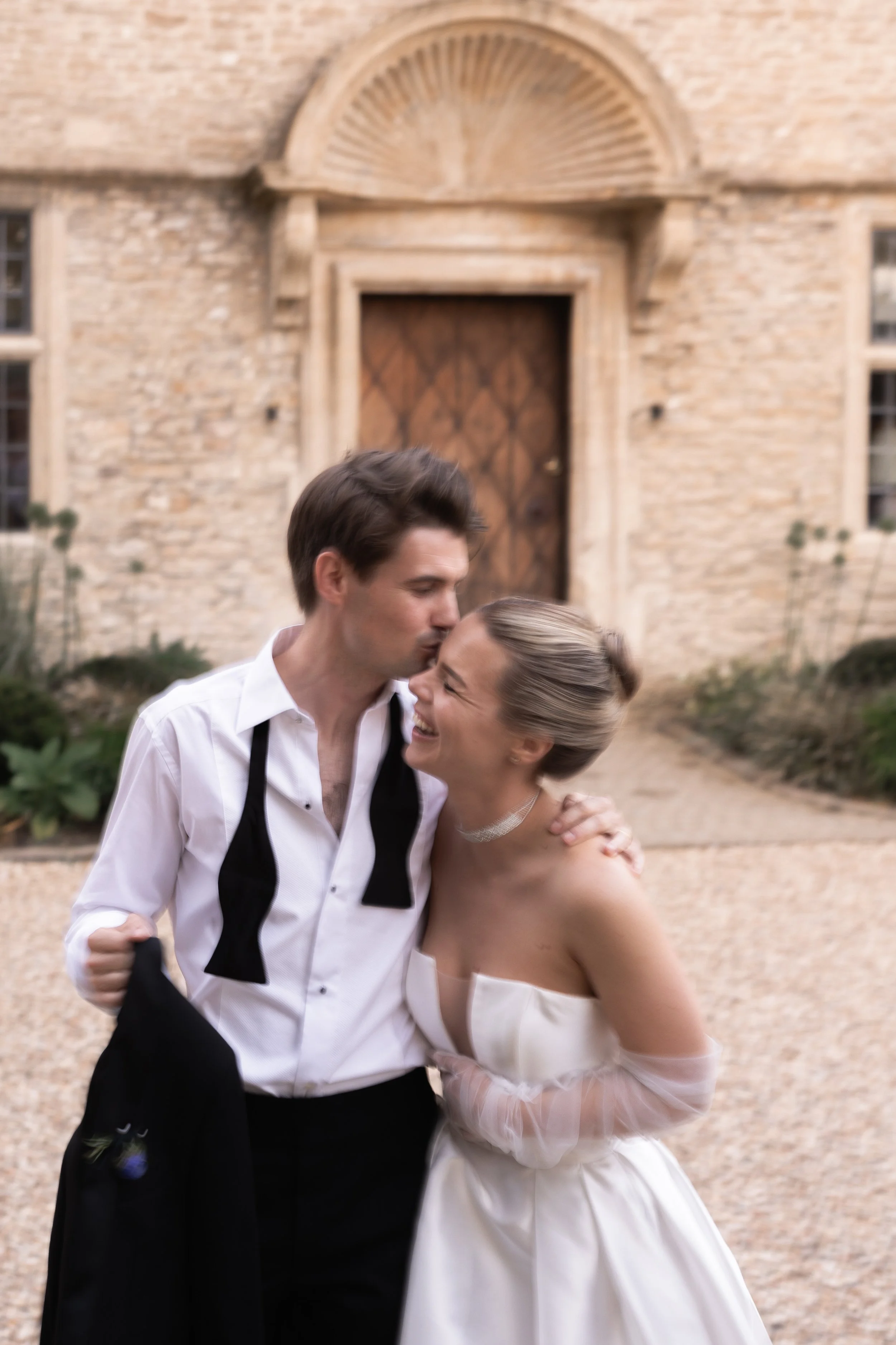 A happy couple, dressed in wedding attire, sharing a joyful moment outside a stone building with a wooden door.